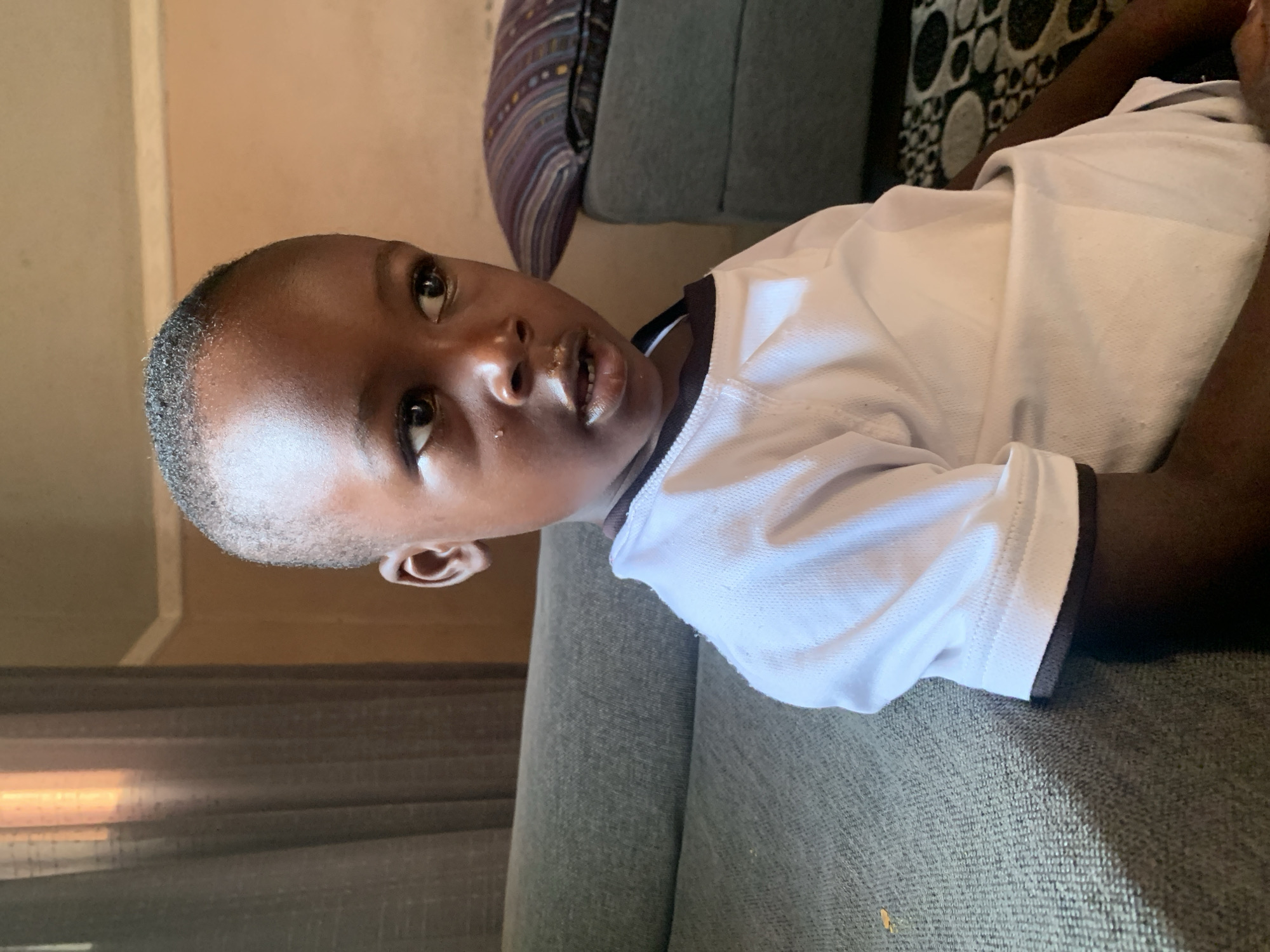 A young Black child with short hair, wearing a white t-shirt with black trim, sits on a gray sofa. The child is looking to their right, with their mouth slightly open. Behind the child, a patterned pillow rests on a dark gray ottoman or seat. A sheer, brown curtain hangs on the left side of the image, with a window or light source visible behind it. The background wall is a neutral, light brown color. The scene appears to be indoors, likely in a residential setting in Sabon Tasha, Nigeria.