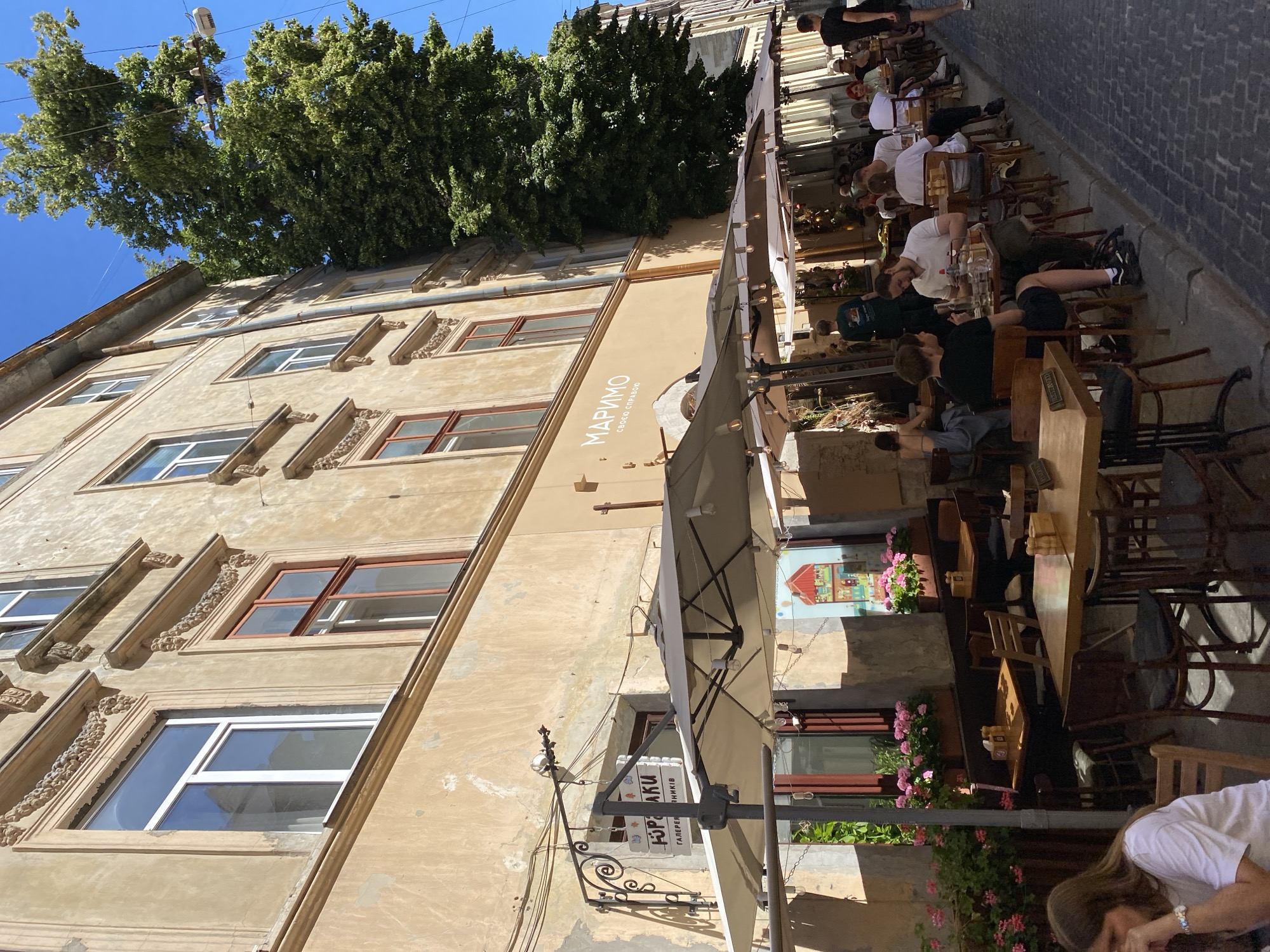 The image depicts an outdoor cafe scene on a sunny day in Lviv, Ukraine. The cafe is situated along a cobblestone street, with tables and chairs set up under white umbrellas, providing shade for patrons. People are seated at various tables, engaged in conversations and enjoying their meals. A woman in the foreground is seen sitting at a table, looking down. The cafe is attached to an old, beige building with ornate window frames. Above the cafe, there are signs, one of which reads "МАРИМО" and another with Cyrillic text. A large, leafy green tree grows beside the building, its branches extending towards the sky. The overall atmosphere suggests a relaxed and pleasant outdoor dining experience.