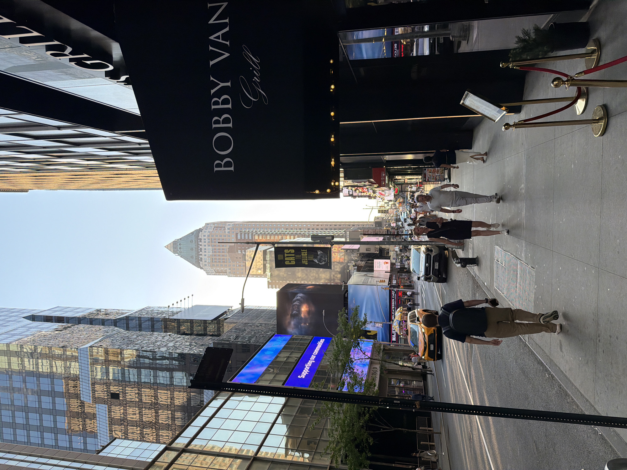 This photograph showcases a scene in New York City, United States, focusing on an urban landscape.
The foreground features "Bobby Van Grill" signage hanging above a sidewalk. The sidewalk is populated with pedestrians. Cars are seen on the street, including a yellow taxi cab.
In the background, skyscrapers dominate the cityscape. Advertisements are visible on the buildings, including one for the Broadway play "Cats."