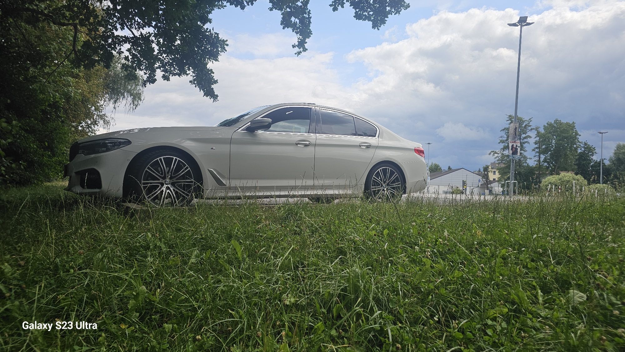 A white sedan is parked on the side of a road in Schleiz, Germany. The car is positioned on the right side of the frame, with its front facing left. The vehicle is mostly obscured by tall green grass in the foreground, which is also slightly out of focus. Behind the car, there is a paved road and a sidewalk. Further in the background, several buildings, trees, and a tall lamppost with posters are visible. The sky is cloudy, with patches of blue peeking through. The overall scene suggests an outdoor setting in a suburban or semi-rural area.