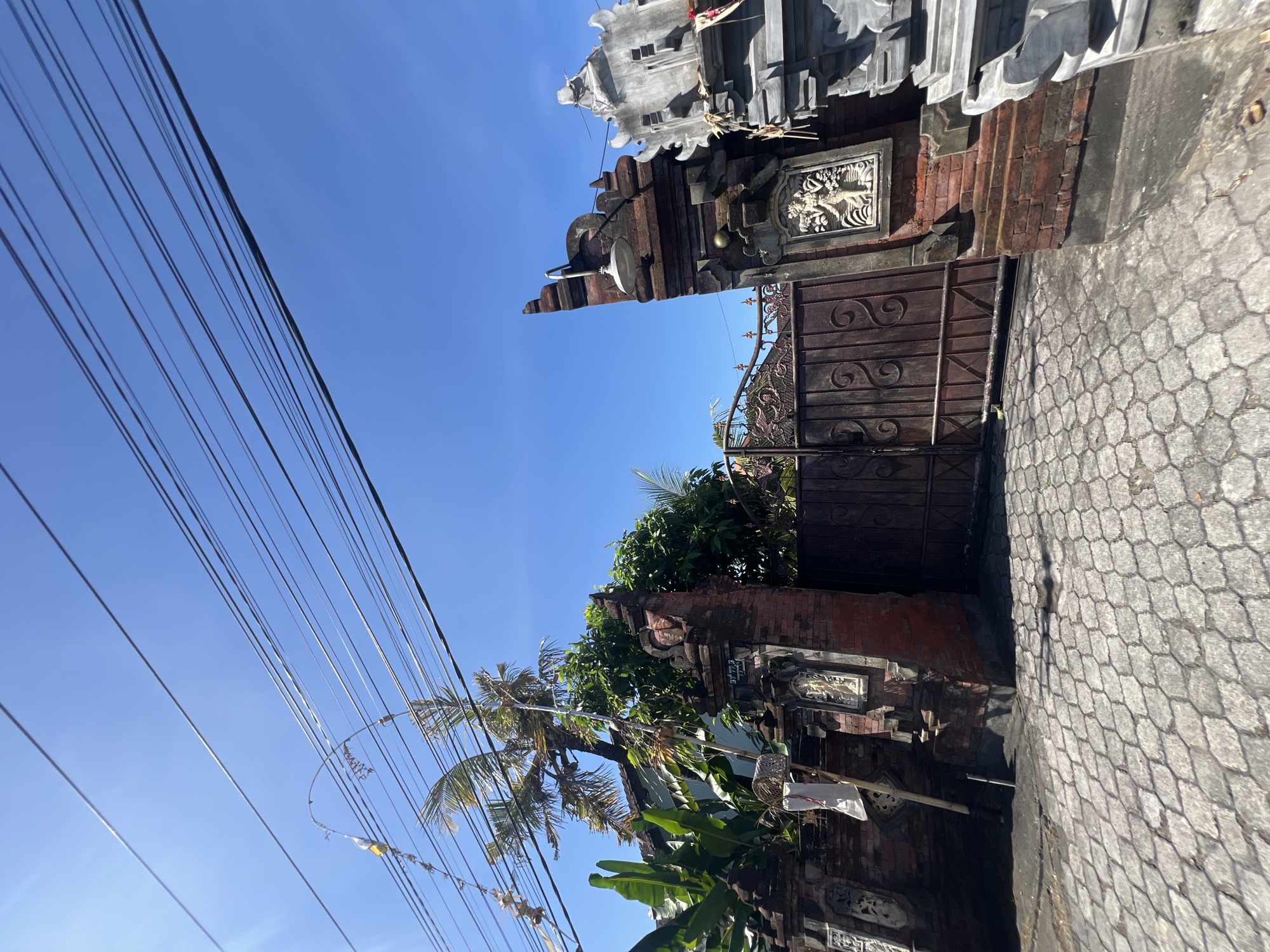 The image shows a traditional Balinese gate in Sukawati, Indonesia. The gate is made of red brick and features ornate carvings of figures on the pillars. A large, dark wooden gate stands behind the brick pillars. Banana trees and a palm tree are visible behind the gate, along with the roof of a building. Overhead, numerous electrical wires crisscross against a clear blue sky. The ground in front of the gate is paved with interlocking hexagonal stones. The overall scene suggests a sunny day in a tropical environment, possibly a residential area or a temple.
