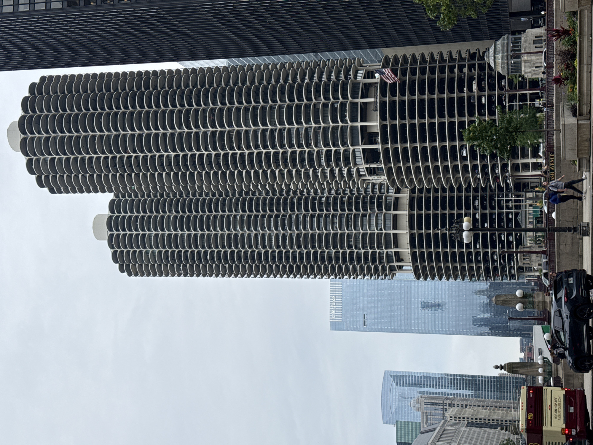 This media file captures an exterior, medium-angle shot of the Marina City towers in Chicago, Illinois. The iconic, corncob-shaped residential skyscrapers dominate the left side of the frame, with their distinctive curved balconies and numerous windows. An American flag is visible on the upper portion of the left tower. To the right of the towers, modern glass skyscrapers rise in the background, suggesting an urban setting.
In the foreground, on the right side of the image, a sidewalk is visible with a few people walking. One person is wearing a blue top and dark pants, while another is wearing a gray top and dark pants. A black SUV is parked on the street, and a red and white double-decker bus is also visible, with visible text on its side: "HOP-ON HOP-OFF" and "BIRDSONGBUS.COM". Streetlights with multiple globes are positioned along the sidewalk.
The overall lighting suggests it is daytime, and the sky appears overcast, indicating potentially cloudy weather. The scene depicts a busy urban environment in Chicago, with a mix of architectural styles and the presence of transportation, suggesting a typical day in the city.