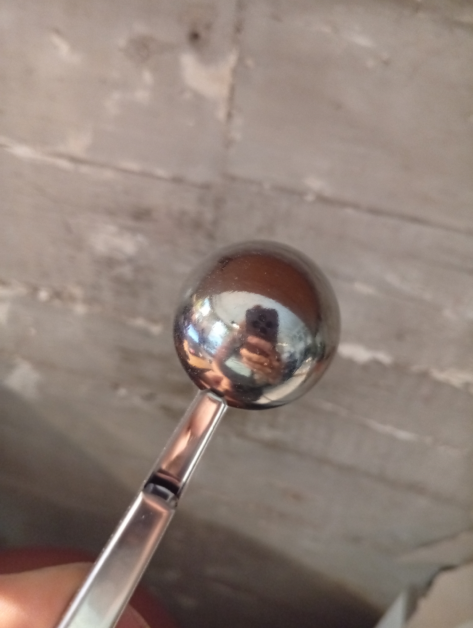 A close-up photograph shows a chrome-plated spherical object attached to a slender, square-profiled handle. The spherical object has a highly reflective surface, mirroring the surrounding environment and a person holding a smartphone. The handle features a small rectangular hole near its attachment point to the sphere. The background is a blurry, mottled gray surface. There is no indication of the location in the city of Buenos Aires, Argentina.