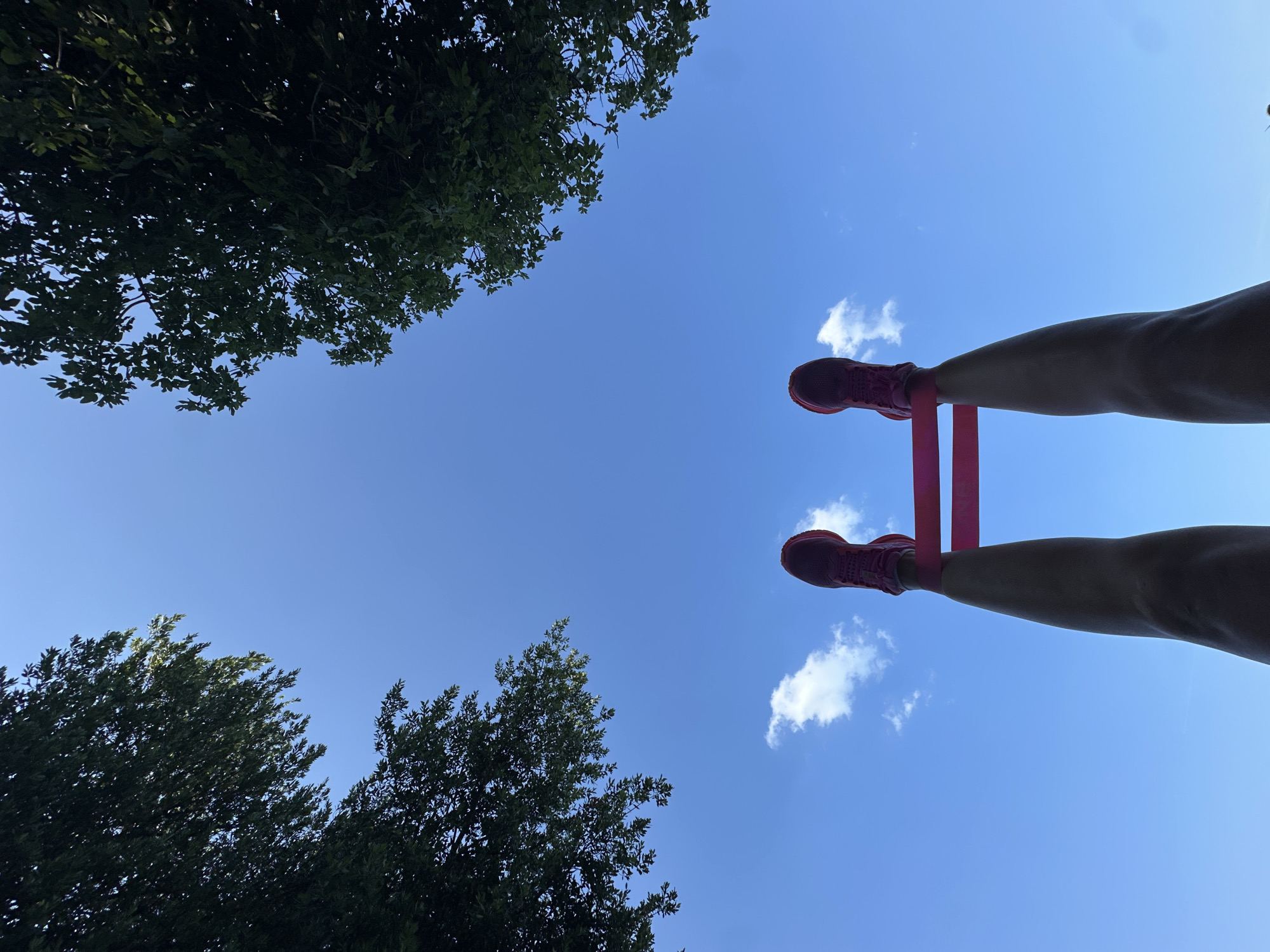 The image shows a low-angle shot of a person's legs, wearing pink athletic shoes and a pink resistance band around their ankles, extended upwards against a bright blue sky with scattered white clouds. Two dark green, leafy trees frame the upper corners of the image. The perspective suggests the person might be lying down or in a seated position, looking up at the sky. The location is identified as Boreti, Montenegro, a coastal area known for its natural beauty.