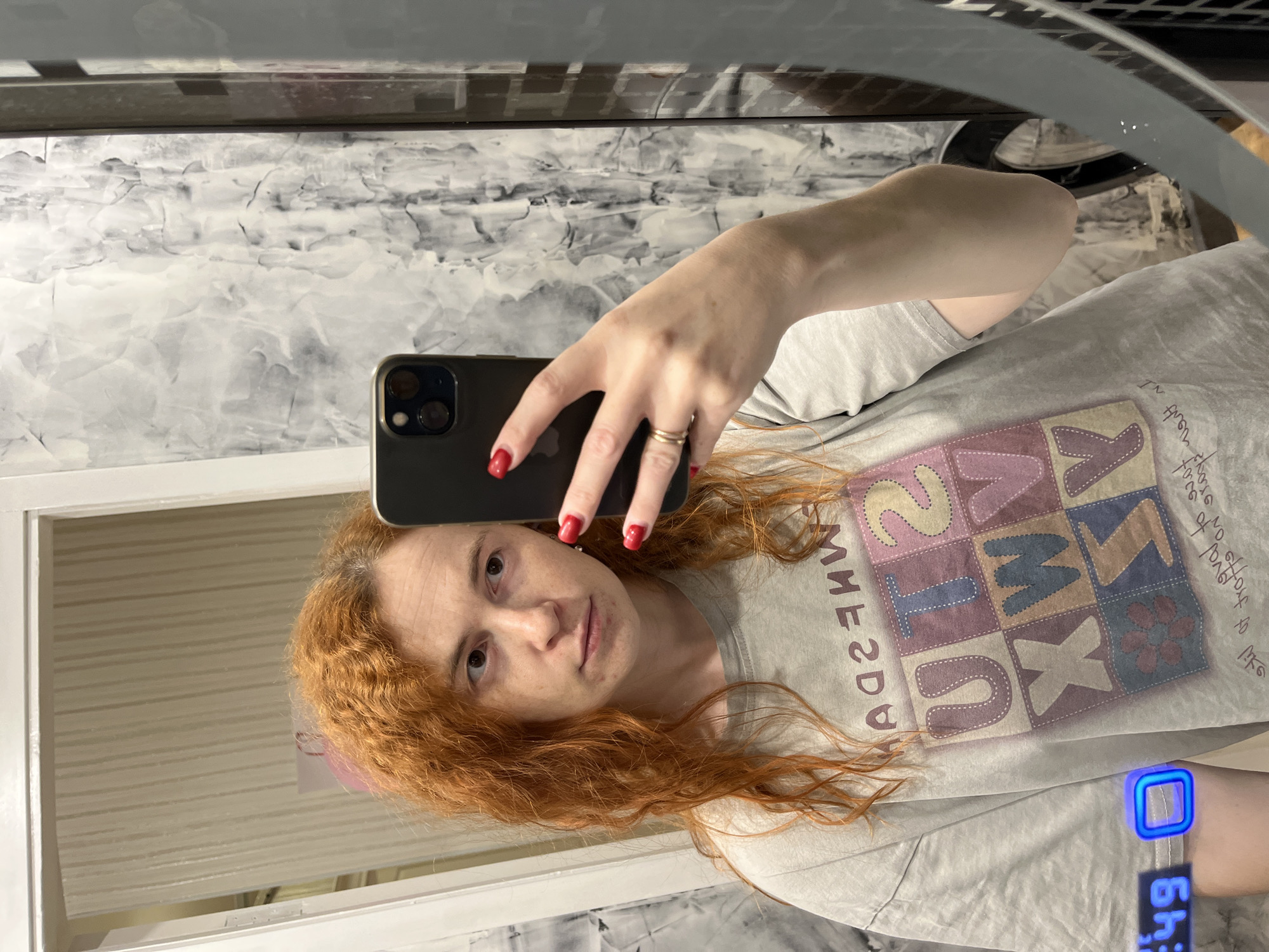 A person with long, red, curly hair takes a mirror selfie with a black smartphone. The person is wearing a light grey t-shirt with a large graphic featuring a patchwork of letters and designs. The time 6:49 is displayed on a blue digital device on the person's wrist. The background includes a marbled grey and white wall and a white door frame. The location appears to be indoors, possibly a bathroom. The city and country are Chaikovsky, Russia.