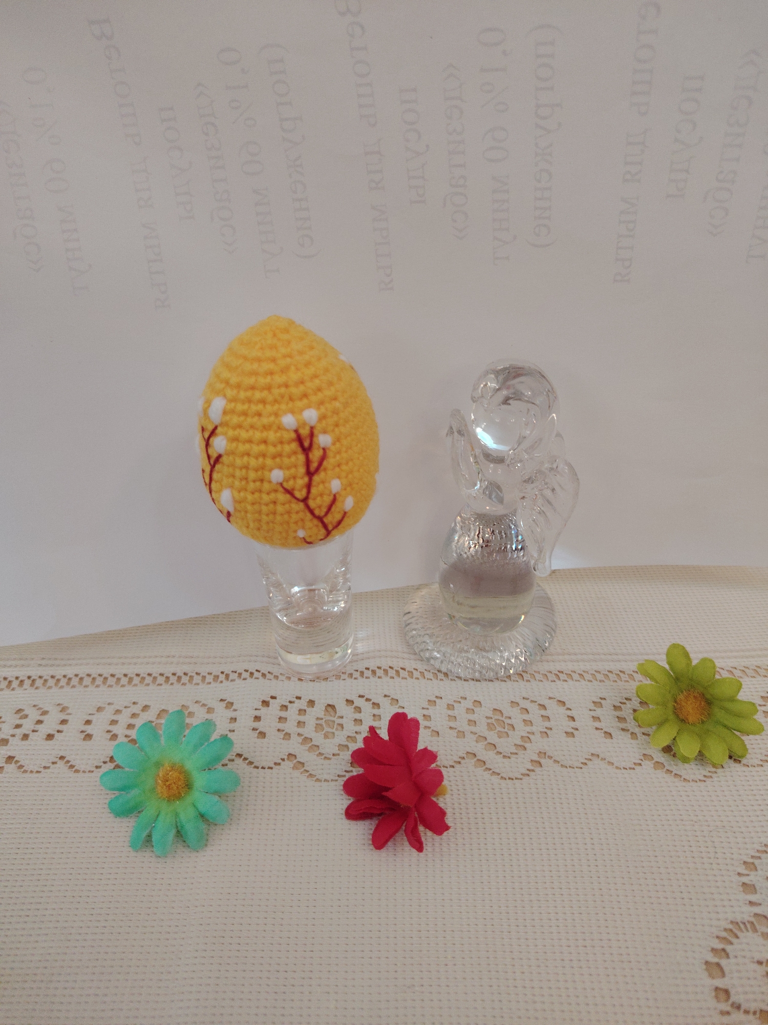 On a lace-edged, off-white tablecloth, a decorative scene is arranged. A yellow, crocheted egg decorated with brown branches and white blossoms sits inside a clear shot glass. Adjacent to the egg is a glass angel figurine. Three artificial flowers—one turquoise, one red, and one green—are scattered across the tablecloth. The backdrop is a white surface printed with text in an unfamiliar alphabet. The location is Sarapul, Russia.