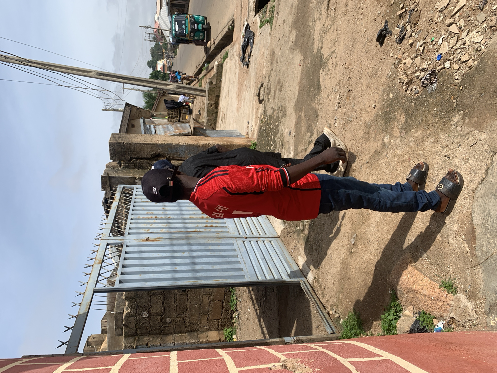 The image depicts a scene on a street in Jos, Nigeria, likely on a sunny day given the clear sky and shadows. In the foreground, a person wearing a red long-sleeved shirt with white trim and dark blue jeans stands facing away from the camera. They are wearing dark sandals. To the left, a brick wall painted red is visible. Behind the person, a light blue metal gate with pointed spikes on top is partially open, revealing a courtyard or enclosed area with a brick wall and some sparse vegetation. Further down the street, a bustling scene unfolds with several people, a green auto-rickshaw, and various buildings lining the road. The auto-rickshaw has "GIFT FROM GOD" written on its roof and license plate "B18". Power lines are visible overhead, supported by a concrete pole. The ground is a mixture of dirt and concrete, with some scattered debris. The overall impression is of a busy urban environment in Nigeria.