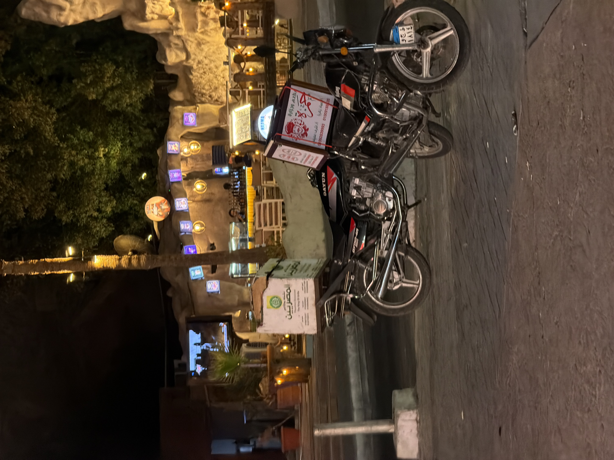 The image depicts a nighttime scene on a paved street in Sharm El Sheikh, Egypt. Two motorcycles with attached food delivery boxes are parked on the street. The motorcycle in the foreground has a white box with Arabic writing and a logo on its rear. The motorcycle behind it has a black box with red trim and similar Arabic writing and logos. Behind the motorcycles, a restaurant or cafe is visible with outdoor seating, lights, and trees. The overall environment suggests a commercial or entertainment area.