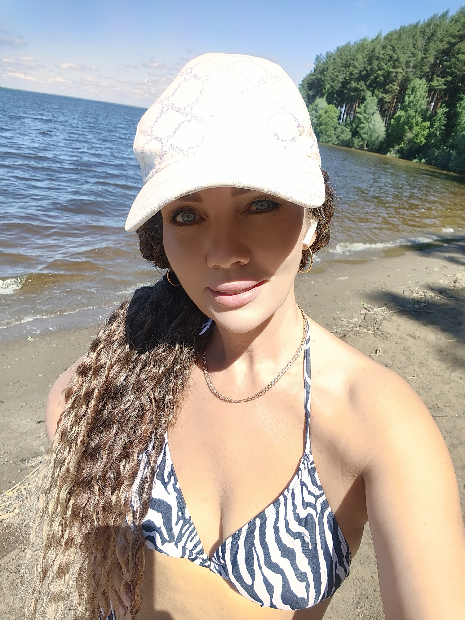 A woman wearing a white baseball cap and a black and white zebra-print bikini top is standing on a sandy beach. Her long, curly, blonde and brown hair is to her left. She is wearing a gold chain necklace and gold hoop earrings. The water of a lake is behind her, and trees are visible in the distance. The sky is blue with some clouds. The scene appears to be in a natural outdoor environment.