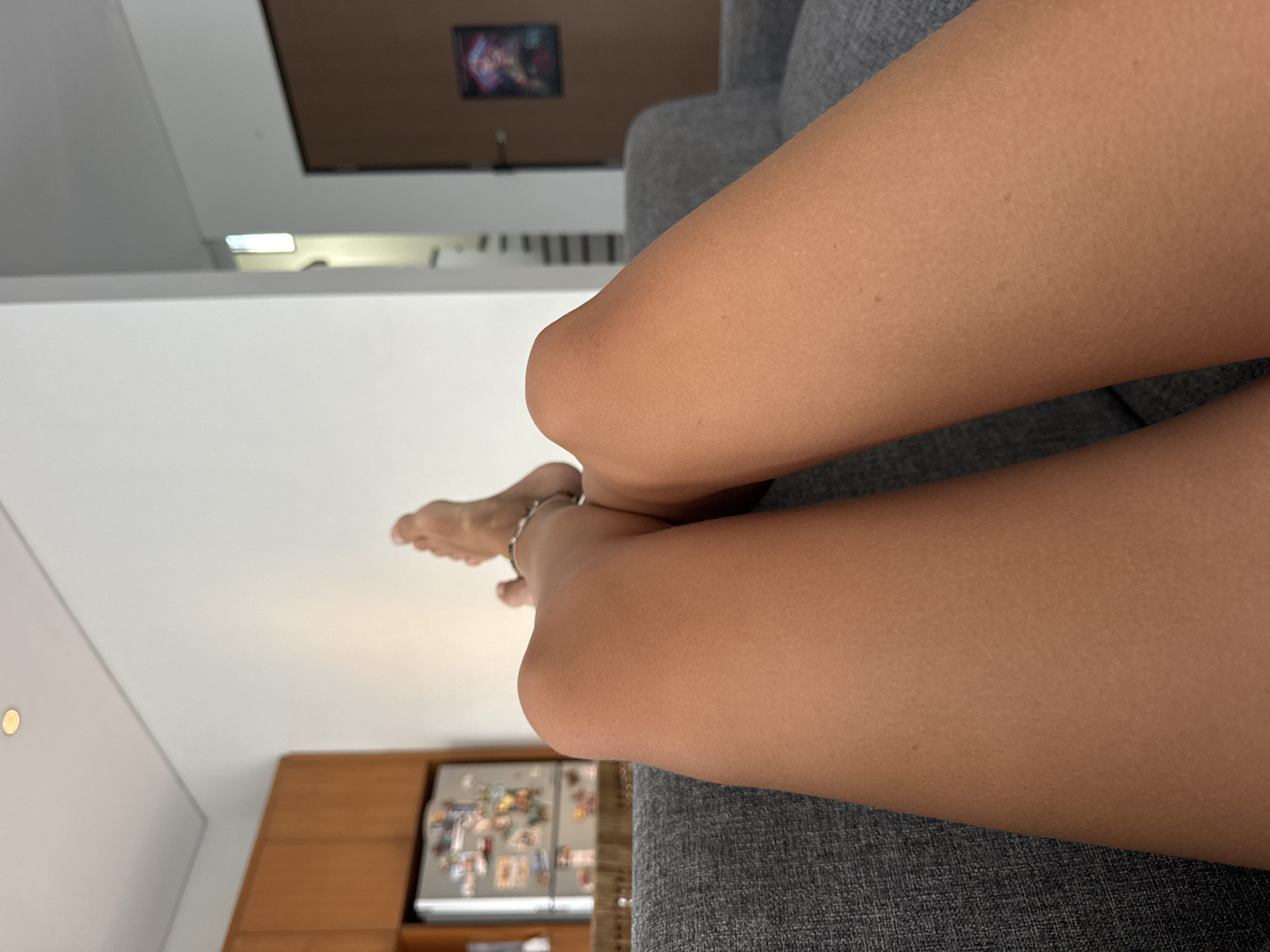 The image shows a close-up view of a person's legs resting on a gray sofa. The legs are tanned and slightly bent. A silver anklet is visible on one ankle. The background includes a white wall, a brown cabinet, and a refrigerator with magnets. The location is a room inside a building.