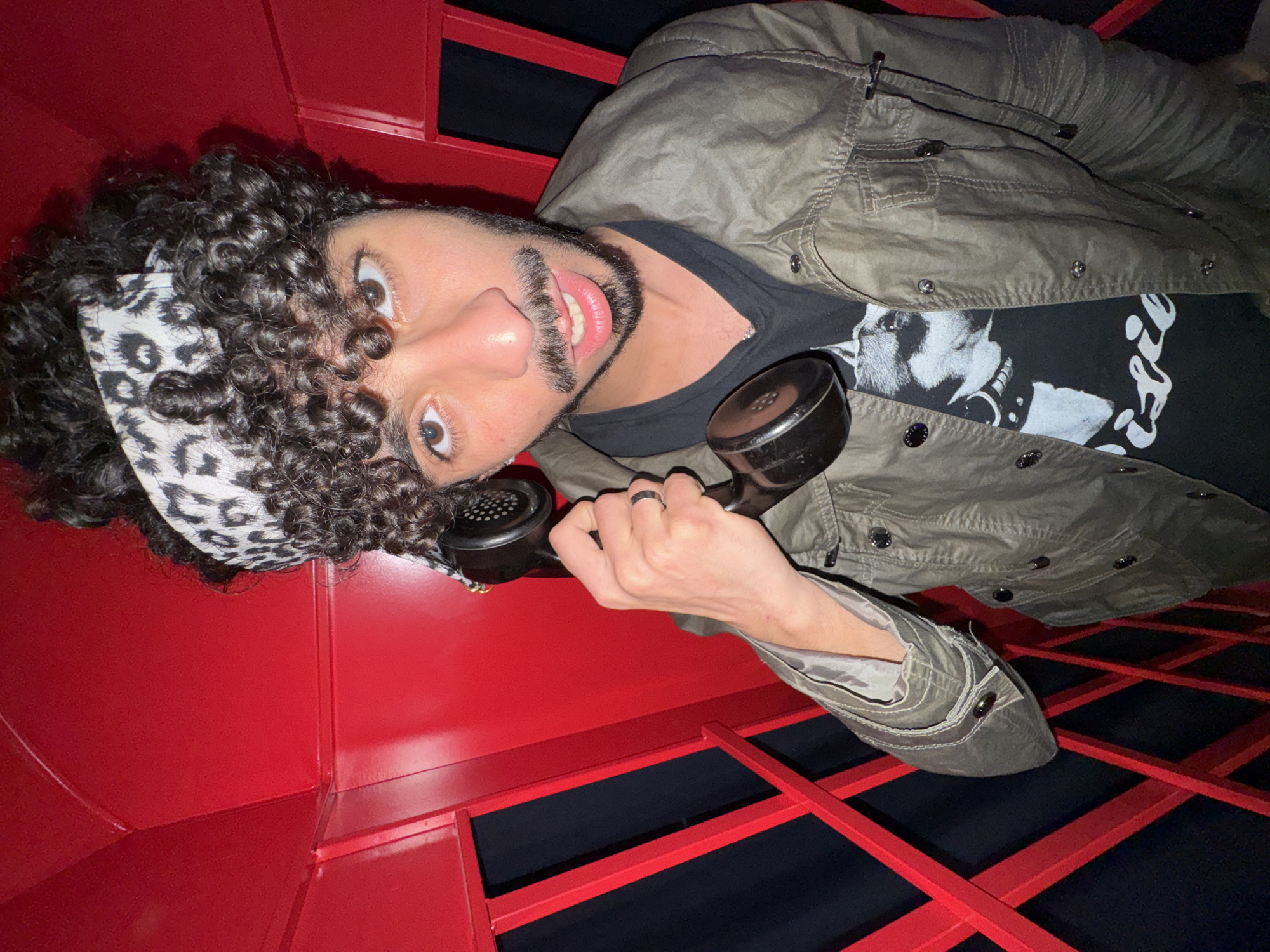 A man with curly hair, a bandana, and a beard is holding a black rotary phone in what appears to be a red telephone booth. He is wearing a gray jacket over a black t-shirt with a white graphic. The booth has a black and red grid-like structure. The photograph was taken in Créteil, France.