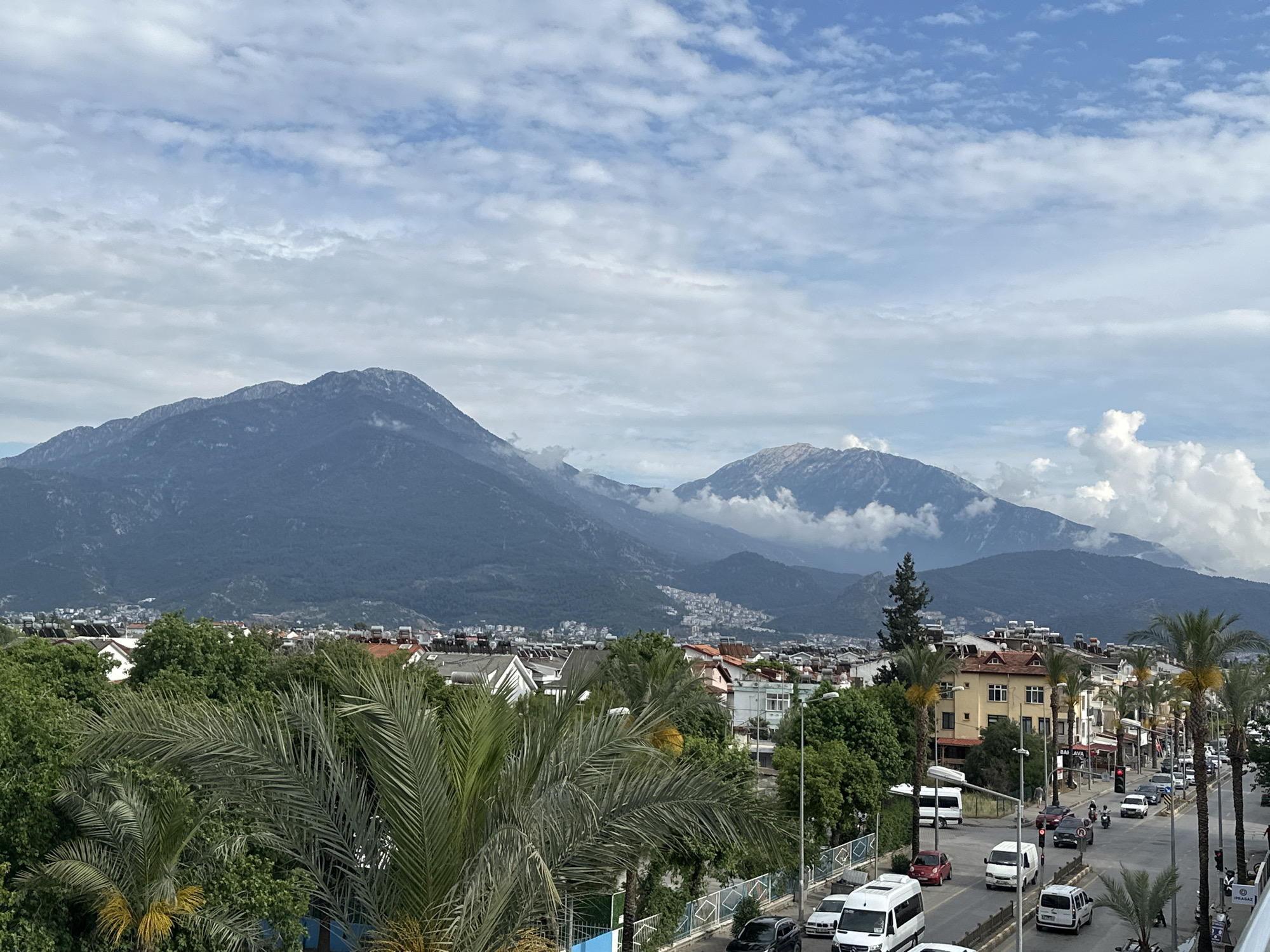 The image presents a cityscape in Fethiye, Turkey, under a sky patterned with scattered white clouds against a blue expanse. In the background, large mountains rise, partially obscured by clouds. The midground features buildings with red-tiled roofs interspersed with green trees, providing a buffer between the urban area and the mountains. In the foreground, a street scene unfolds with moderate traffic consisting of cars, vans, and a motorcycle. Palm trees line the street, adding to the tropical ambiance. The buildings adjacent to the street appear to be a mix of residential and commercial properties. The image is captured from an elevated viewpoint, possibly a building or hill overlooking the city.