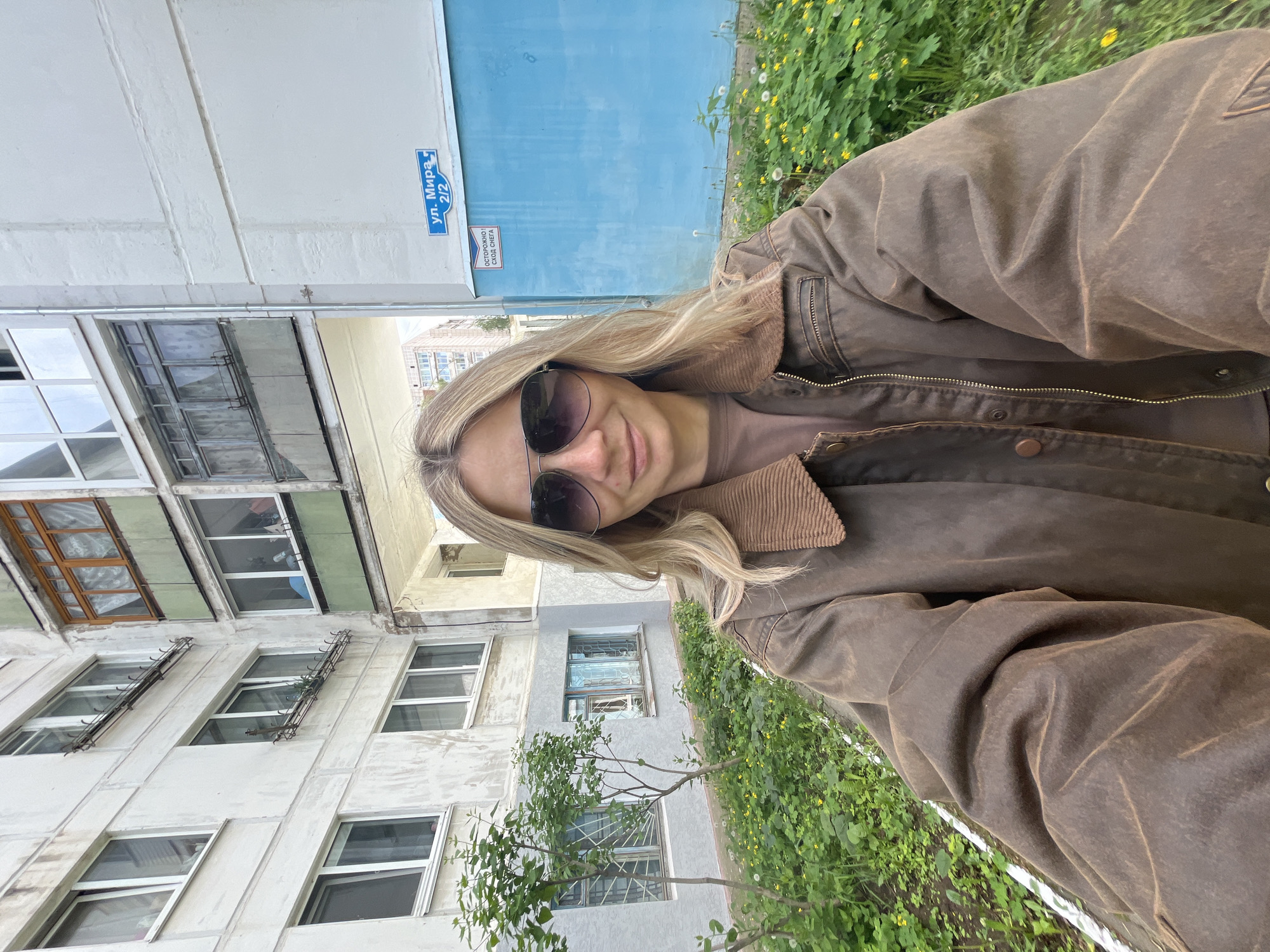A woman with blond hair and sunglasses is taking a selfie outdoors. She is wearing a brown jacket and a brown t-shirt. Behind her is a building with multiple windows. There is green foliage and yellow flowers to her right, and a blue door and a street sign that reads "ул. Мира 2/2", which means "Mira Street 2/2", on the building. Based on the street sign and architecture, the location is likely in the city of Chaikovsky, Russia.