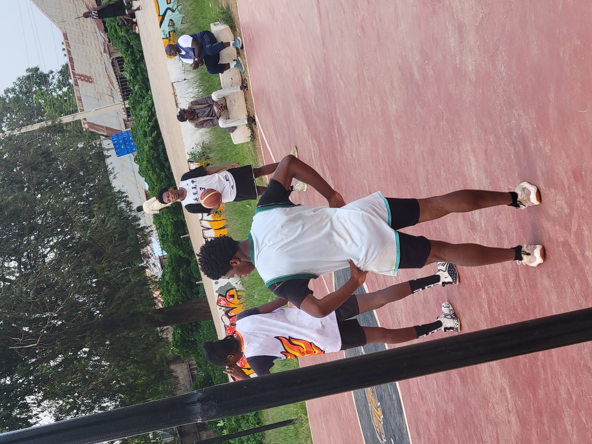 An outdoor basketball court with a red surface is captured in this medium shot. In the foreground, a young Black male is standing with his back to the camera, wearing a white sleeveless jersey with teal trim and black shorts. He is positioned on the right side of the court. To his left, a young Black male is facing the camera, holding a basketball, and wearing a white jersey with black trim and Chinese characters, along with black shorts and black and white sneakers. Behind him, another young Black male is seen from the back, wearing a white jersey with orange flame graphics and black shorts. Further in the background, to the right of the court, two adult Black males are seated on a concrete ledge. One is wearing a blue shirt and dark pants, and the other is wearing a grey jacket and light-colored pants. The environment includes a large tree with dark green foliage to the left, and a line of buildings with corrugated metal roofs in the distance. The sky is overcast. The scene appears to be a casual basketball game or practice.