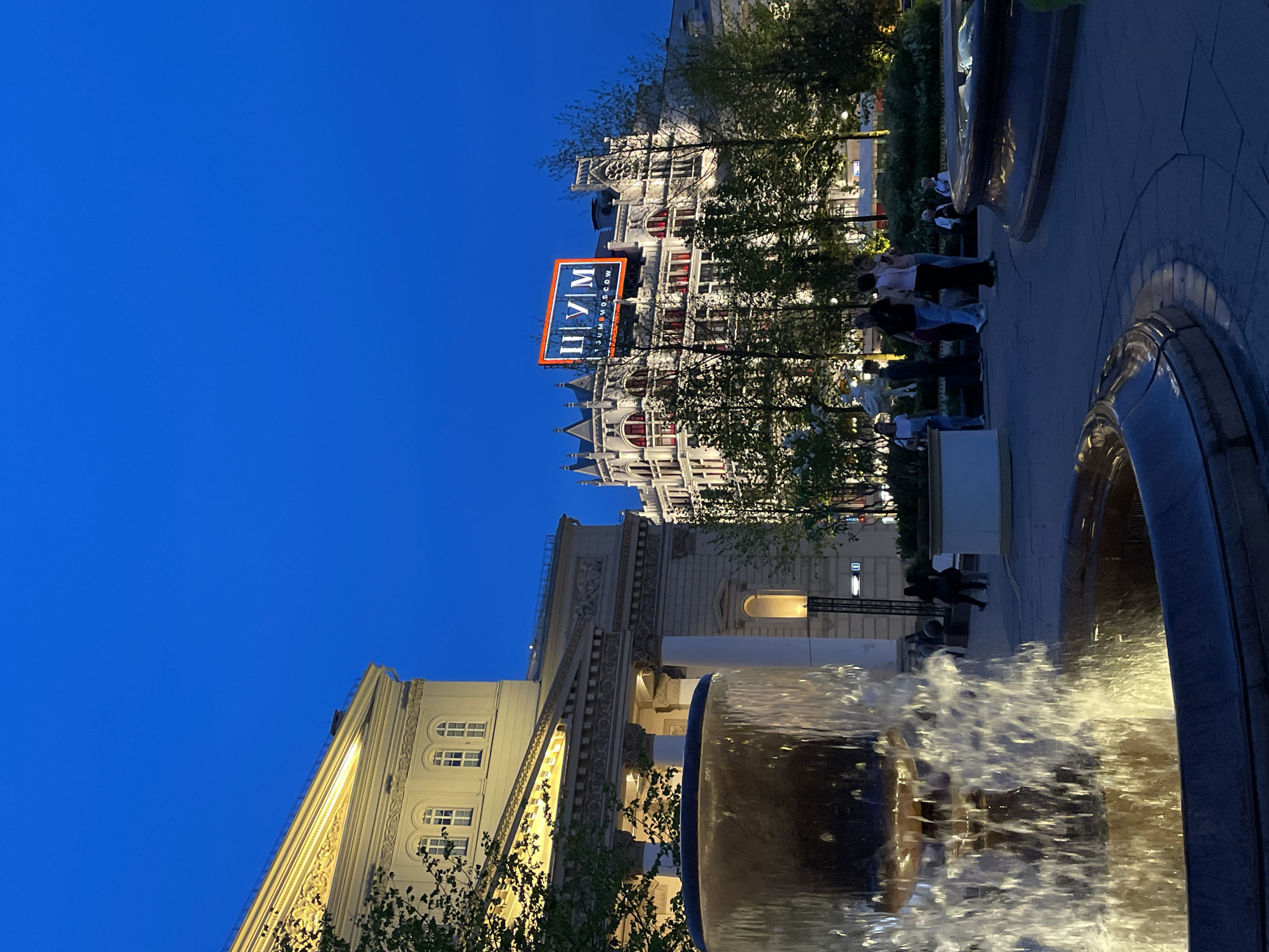 The image shows a nighttime view of the GUM department store in Moscow, Russia, with a water fountain in the foreground. The sky is deep blue. The building is illuminated, and the "ГУМ" sign is prominently displayed. A group of people are gathered on the sidewalk near the building. The fountain is also lit, and the water is in motion.