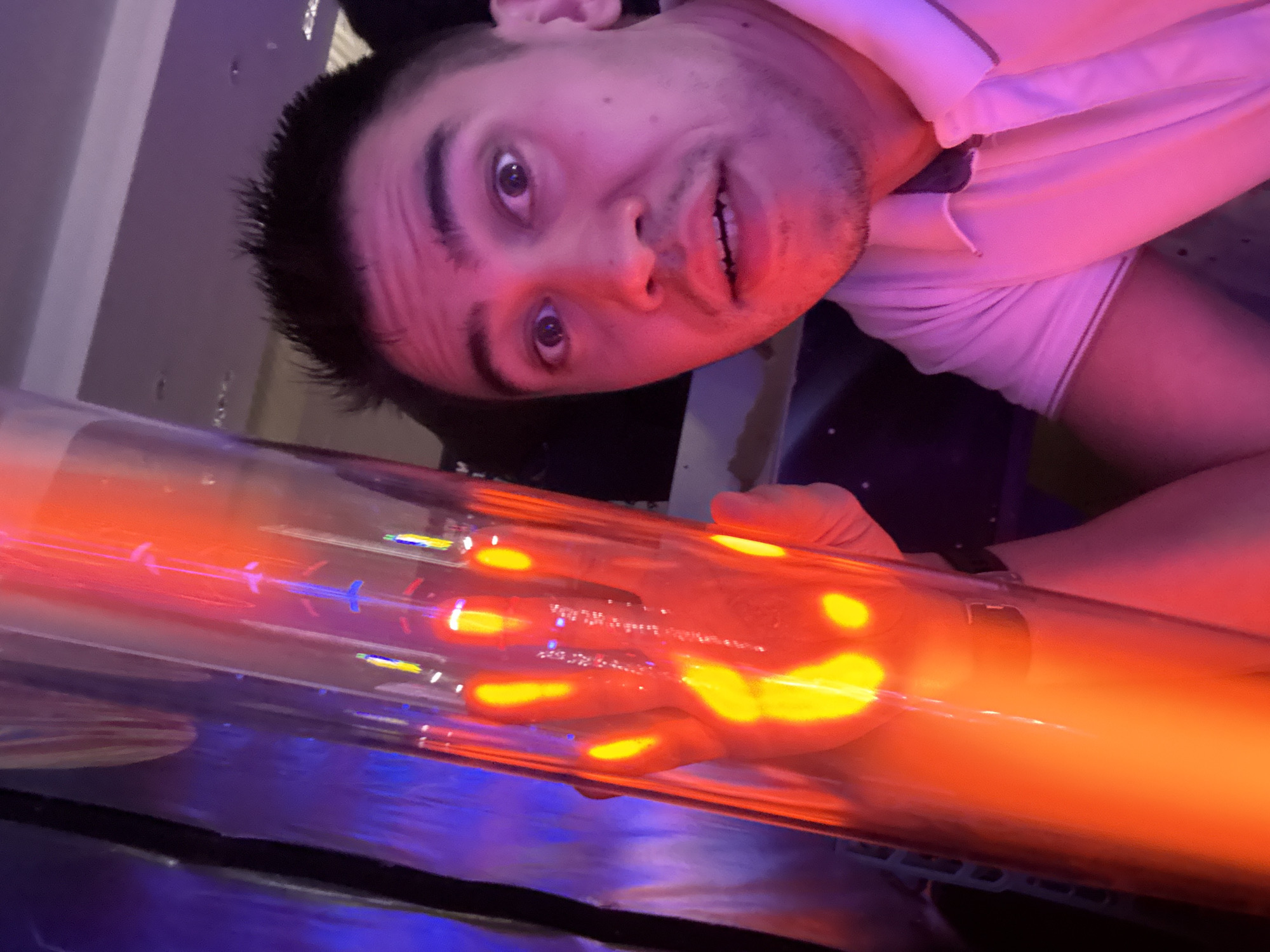 A man is holding a clear tube that is glowing orange and yellow. He appears to be indoors, possibly in an entertainment venue in Kazan, Russia. The lighting in the area is mixed, with pink, red, and purple hues. The man has dark hair and is wearing a white collared shirt. His expression is somewhat surprised. The environment includes reflective surfaces and what appears to be part of a seating arrangement with dark coloring.