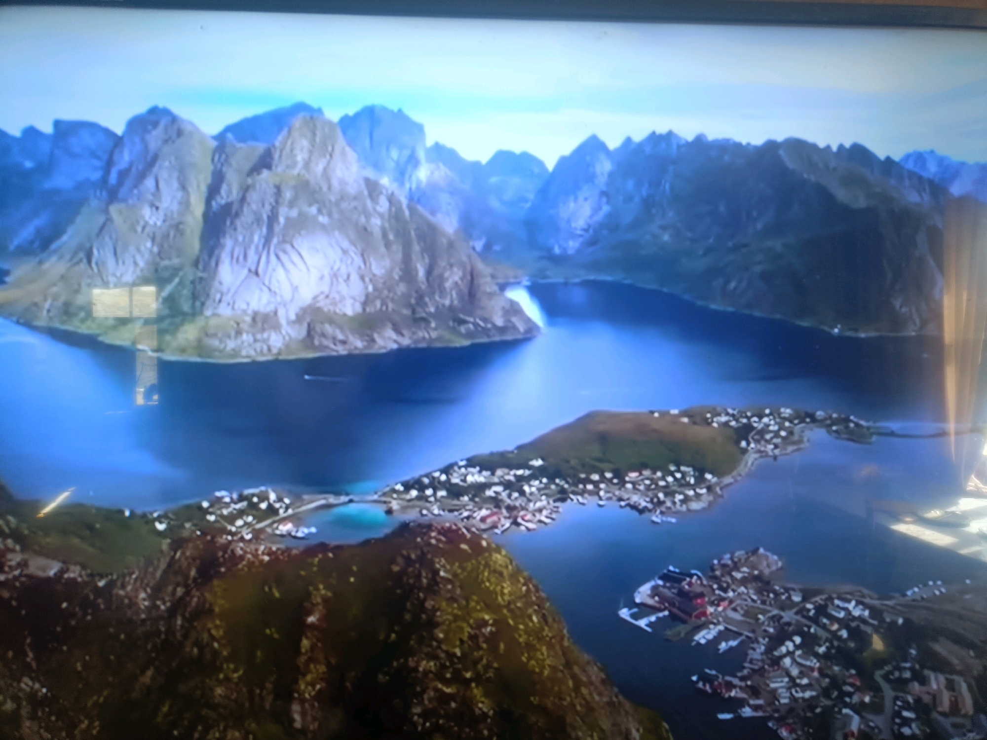 The image is an aerial view of a coastal village nestled among dramatic, jagged mountains. The water is a deep, reflective blue, and the sky is a pale, hazy blue. The village consists of numerous small, white buildings clustered along the coastline, with a harbor filled with boats. In the foreground, a steep, rocky mountain covered in patchy green vegetation rises from the water. The overall impression is one of a remote and rugged natural beauty. There is no indication that the location is in the city of Қасымбек датқа, Kazakhstan.