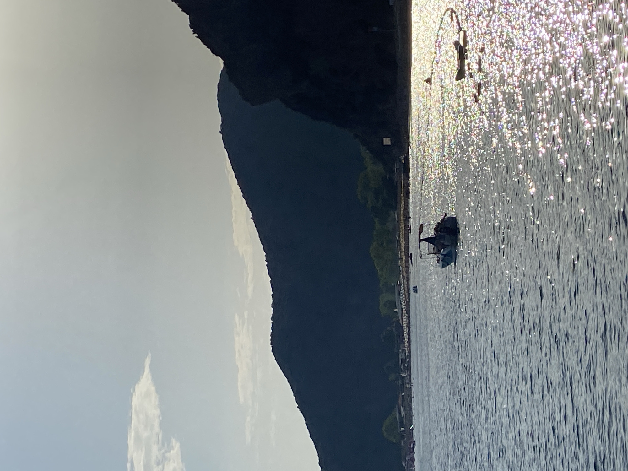 The image shows a coastal scene in Prijevor, Montenegro, under a bright, overcast sky. The foreground is dominated by the calm, dark blue water of the sea, reflecting the sunlight in a shimmering pattern. A small motorboat with a canopy is visible in the middle of the water. In the background, a shoreline is lined with buildings and lush green trees. Beyond the shoreline, steep, dark green mountains rise, casting a shadow over the landscape. A few people are visible on the beach and in the water, suggesting a recreational setting. The overall impression is one of a serene coastal environment during daylight hours.