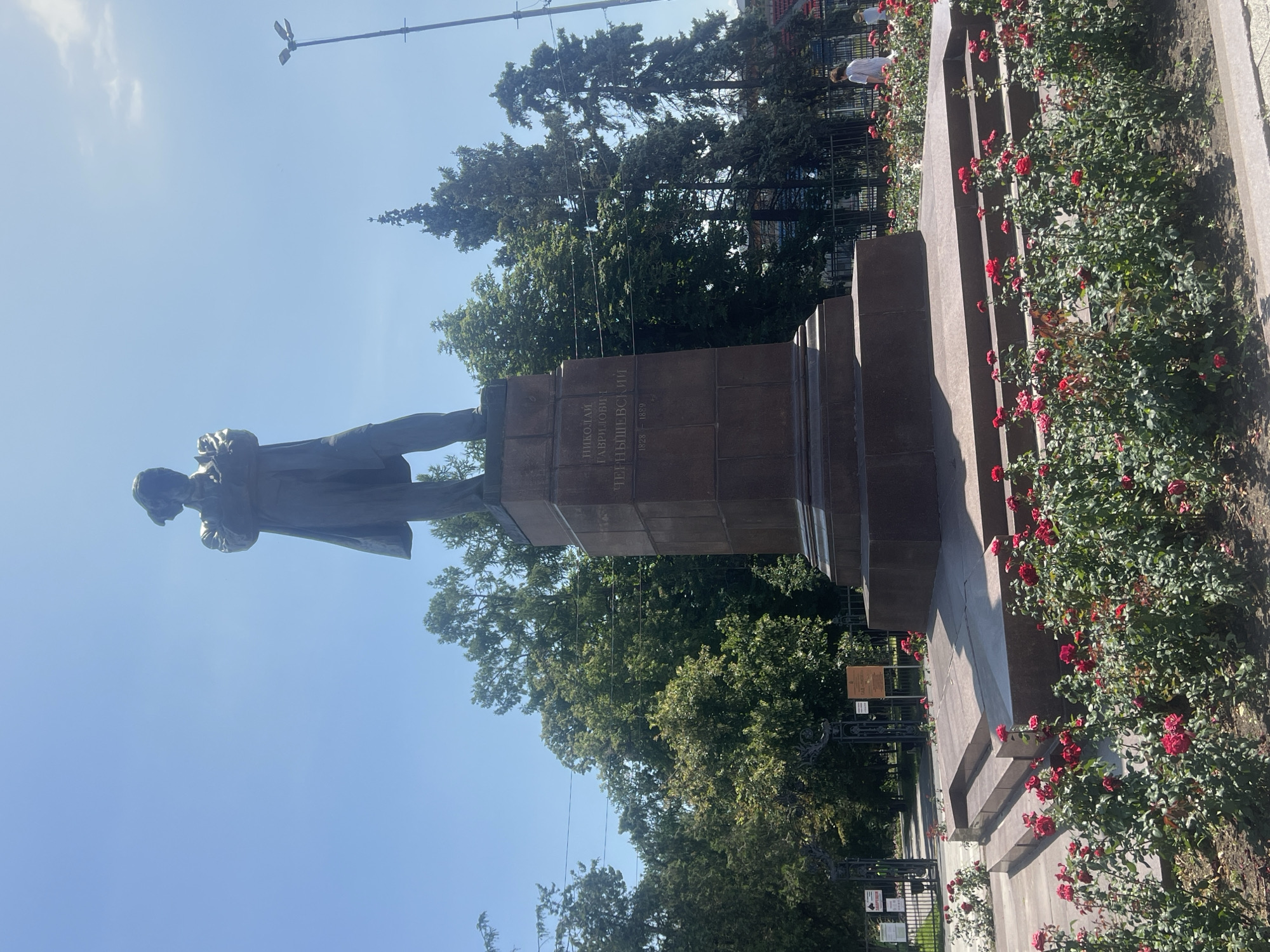 A full shot shows a bronze statue of a man, Nikolai Gavrilovich Chernyshevsky, on a tall granite pedestal. The statue depicts Chernyshevsky standing with his arms crossed and his head tilted slightly, looking to his right. He is dressed in a long coat and trousers. The pedestal is inscribed with his name and dates, 1828-1889.
The statue is situated in a park-like setting, with lush green trees in the background. In the foreground, a bed of vibrant red roses blooms, their petals bright against the dark soil. The sky above is a clear, bright blue, indicating a sunny day. A black wrought-iron fence and gate are visible to the left of the statue, with a path leading through them into the park. In the far right of the image, a street lamp is visible, and a few people can be seen in the distance, one of them walking away from the camera. The scene appears to be a public monument in a city park.