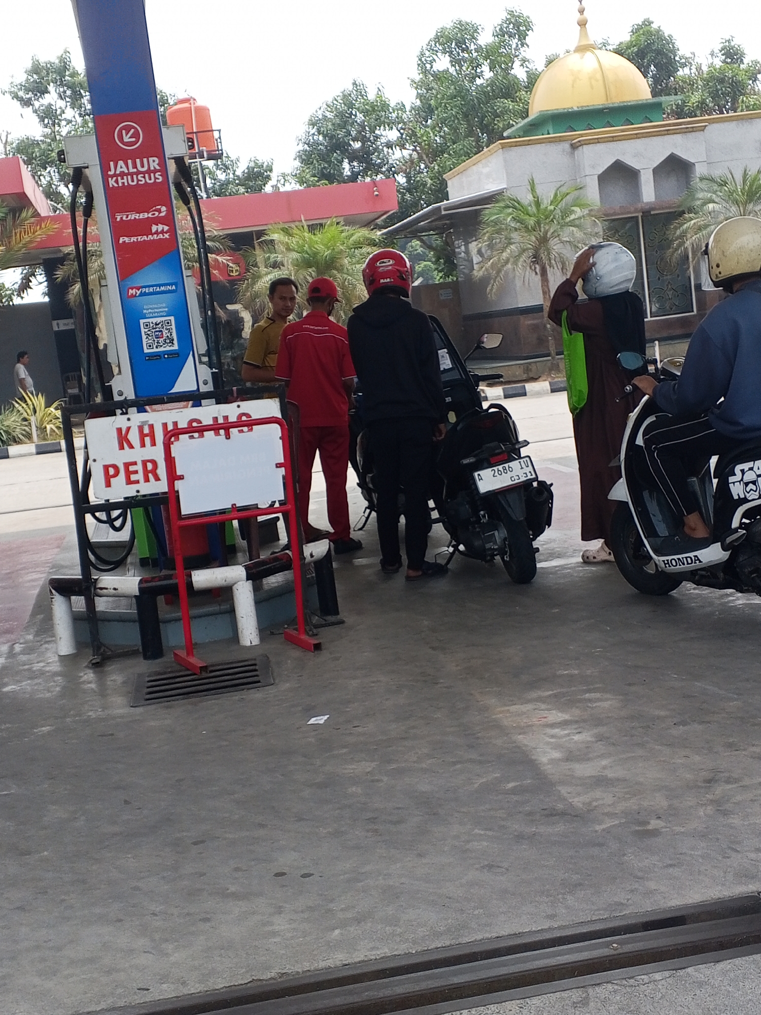 The image depicts a scene at a Pertamina gas station in Ciruas, Indonesia. In the foreground, a red and white sign reads "KHUSUS PERL" (Special Fuel), indicating a restricted lane. A gas pump with the Pertamina logo and "JALUR KHUSUS" (Special Lane) text is visible. Three men, one in a yellow shirt and two in red uniforms, are near the pump. Two motorcycles are parked next to each other, one black with license plate "A 2686 IV" and another white with "HONDA" branding. A woman in a brown robe and hijab, wearing a white helmet, is adjusting her helmet. Another person in a blue shirt, also wearing a yellow helmet, is seated on the white motorcycle. In the background, a building with a gold dome, likely a mosque, is visible, surrounded by trees and palm trees. The ground is paved concrete, and a metal drain grate is present. The lighting suggests it is daytime, possibly under cloudy skies.