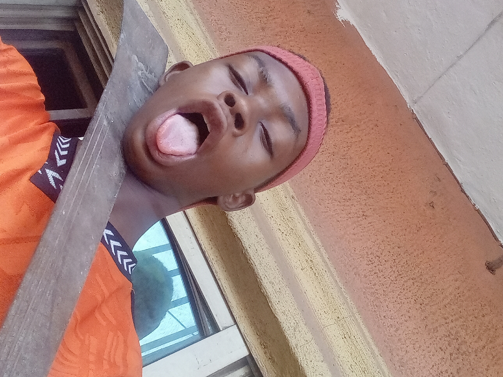 A person wearing a red headband and orange shirt is shown with their eyes closed, mouth open, and tongue sticking out. The person is holding a piece of grey wood to their cheek and neck. The background consists of an orange textured wall, a window, and a piece of white wall. The location is most likely Nkpor, Nigeria.