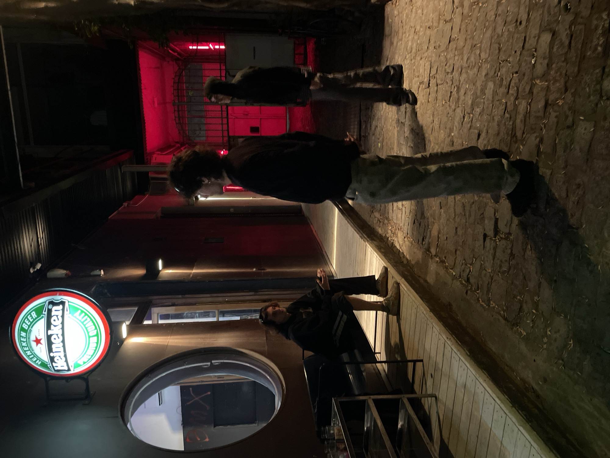 The image displays a nighttime scene within an urban alleyway, likely in Buenos Aires, characterized by atmospheric lighting and several individuals. On the left side, a prominent, circular Heineken Beer sign glows brightly, featuring its iconic green, red, and white logo. The sign clearly reads "HEINEKEN BEER" and "PREMIUM QUALITY." Below it, a dark, circular window or opening is visible on the building facade.
In the foreground, a person with dark, medium-length hair is seated casually on a wooden bench or ledge along the building. They are dressed in a dark hoodie, dark pants, and light-colored boots, appearing relaxed as they look towards the right of the frame. Further into the alley, a second individual stands, also with dark, curly hair, wearing a dark top and patterned trousers. They are upright, facing left, seemingly observing their surroundings. In the background, a third person is partially visible, standing near a structure that is intensely illuminated by a deep red light, casting a strong crimson glow across that section of the alley.
The ground is paved with rough, dark cobblestones or bricks, with scattered leaves contributing to the urban texture. The overall ambiance is dark and moody, punctuated by the artificial light sources and the striking red backlighting, suggesting a casual evening social setting, possibly outside a bar or restaurant.