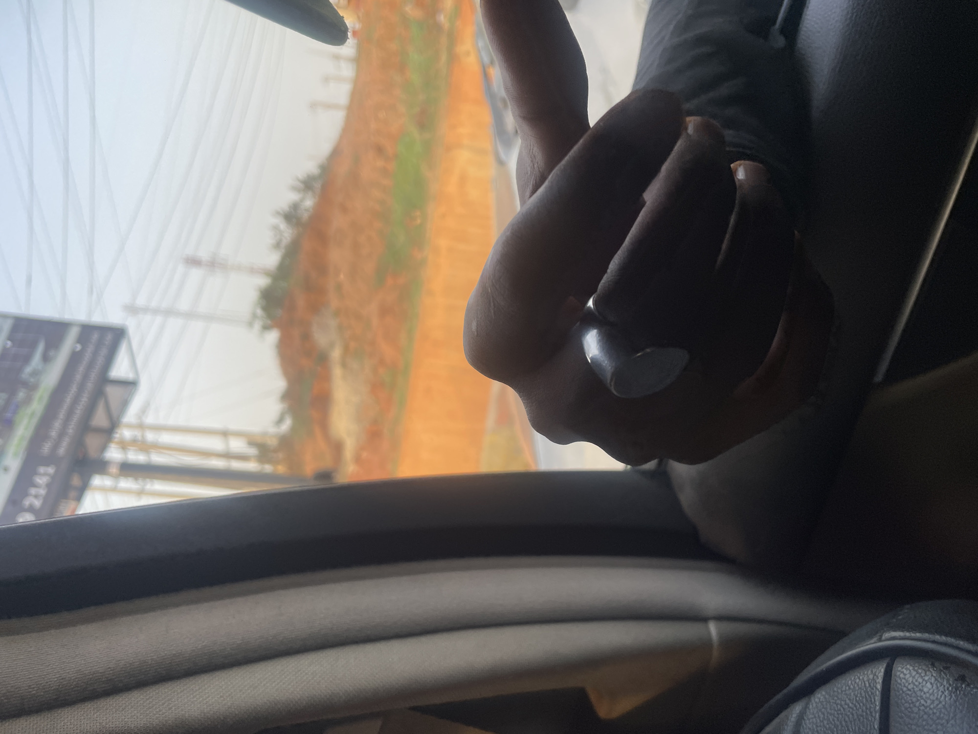 The image presents a view from inside a vehicle, looking out a side window. In the foreground, a person's right hand, with dark skin, is visible, wearing a silver-toned ring on the index finger. The hand appears to be resting casually. Below the hand, elements of the car's interior are visible, including a light gray fabric surface, likely a door panel or armrest, and a darker, possibly leather, surface towards the bottom right.
Outside the car window, the scene is dominated by a hazy, bright sky. Numerous thin lines, characteristic of power lines or utility cables, are stretched across the upper left portion of the view. The landscape outside features a large expanse of reddish-brown ground, possibly sand or dry earth, with scattered patches of green foliage. In the far distance, faint outlines of structures, possibly construction cranes, can be discerned through the haze.
On the left side of the external view, a part of a billboard or sign is visible. The number "2141" is clearly legible on it, along with other unreadable or partially obscured text, which includes what appears to be a "G" and a "P" in a different section.
The overall impression is that of traveling through an urban or semi-urban area during the daytime, under hazy atmospheric conditions. The landscape and visibility align with the provided context of Abuja, Nigeria.