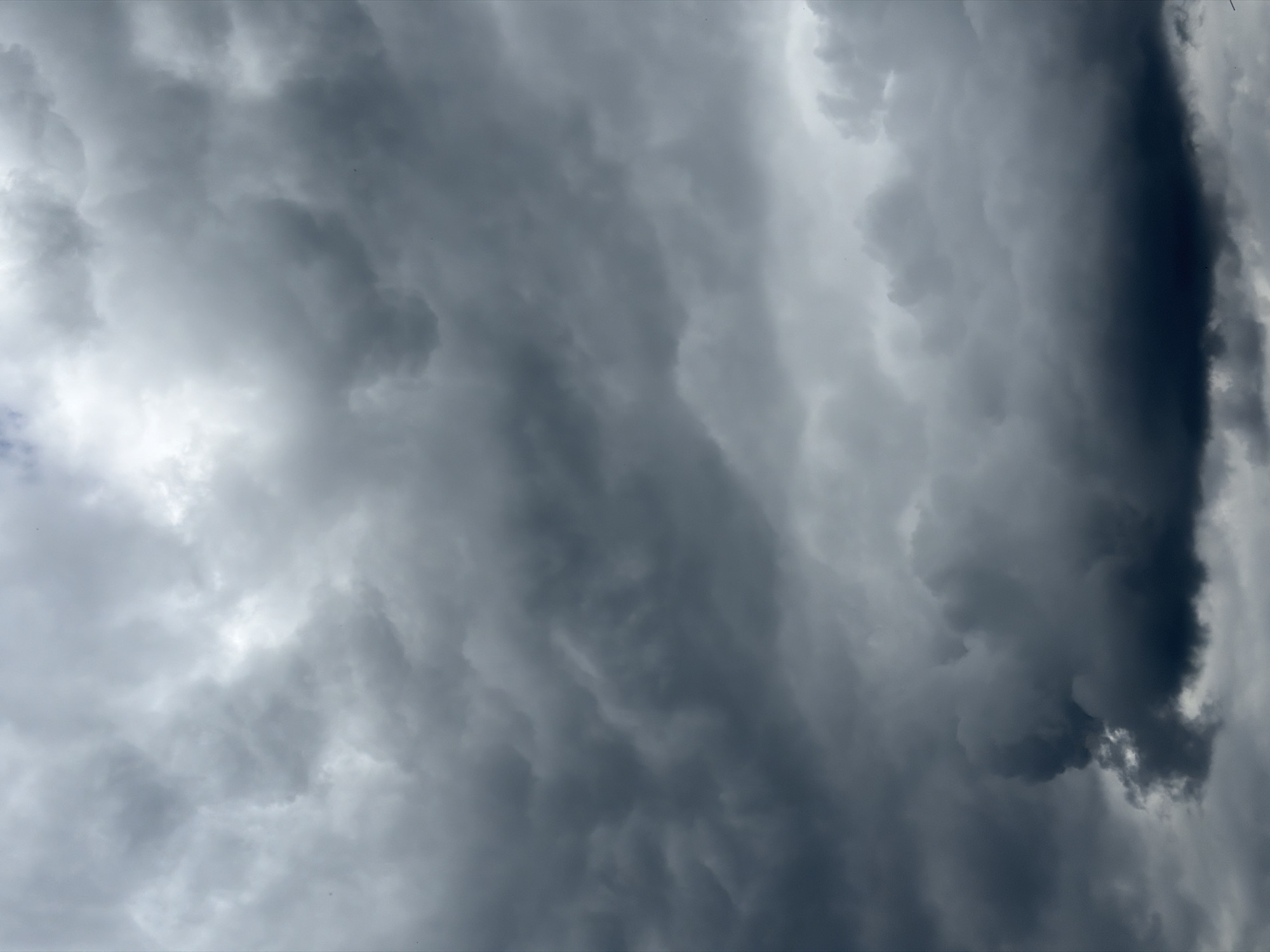 A low-angle, vertical shot captures a sky filled with a dense, turbulent layer of dark gray storm clouds. The clouds are textured and appear to be heavily laden with moisture, with some areas lighter where the sky is still visible, and other areas almost black. The overall impression is one of an impending storm. The image is not specific to any city or identifiable landmarks, and there are no people or objects visible.
