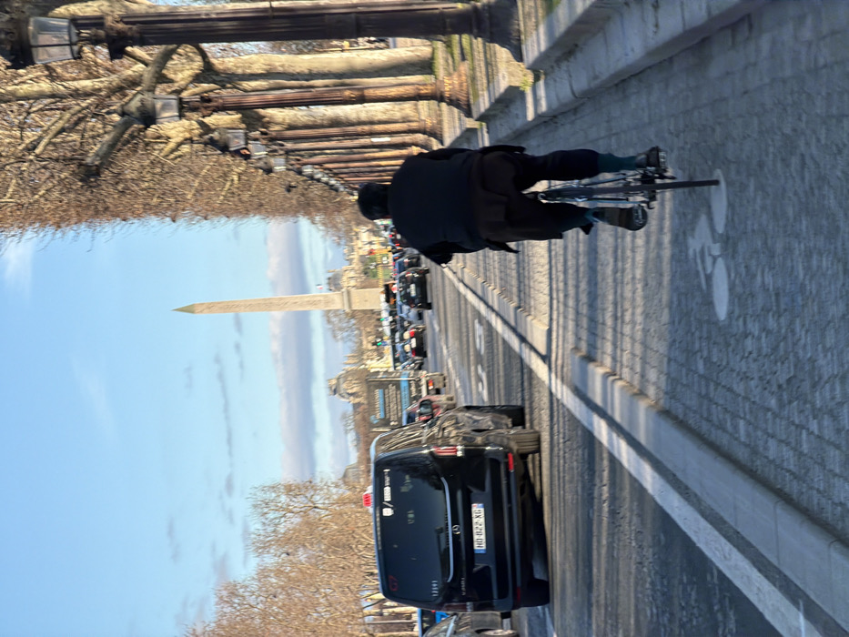 The image displays an urban street scene in Paris, France, captured looking east along the Avenue des Champs-Élysées towards the Place de la Concorde. In the foreground, a person is observed from behind, actively riding a bicycle on a cobblestone cycle path that features a white painted bicycle symbol. The cyclist is dressed in dark attire, comprising a black jacket and dark trousers, with visible dark or greenish-blue socks. This cycle path is delineated from an adjacent multi-lane asphalt road by a low concrete curb.
On the road, vehicle traffic is present and appears congested. The most prominent vehicle is a black Mercedes-Benz V-Class van, equipped with a yellow roof-mounted taxi sign and displaying a French license plate, "HD-822-XG." Other cars and vans are visible in the traffic lanes, exhibiting red taillights. To the right of the cycle path, a linear arrangement of bare deciduous trees stands, interspersed with ornate, multi-fixture lampposts, extending into the distance. The mid-ground is dominated by the Luxor Obelisk, situated centrally within the Place de la Concorde. Beyond the obelisk, classical Parisian architecture, consistent with the Tuileries Garden and Louvre Palace complex, is discernible. The scene is illuminated by daylight under a clear blue sky with scattered white clouds.