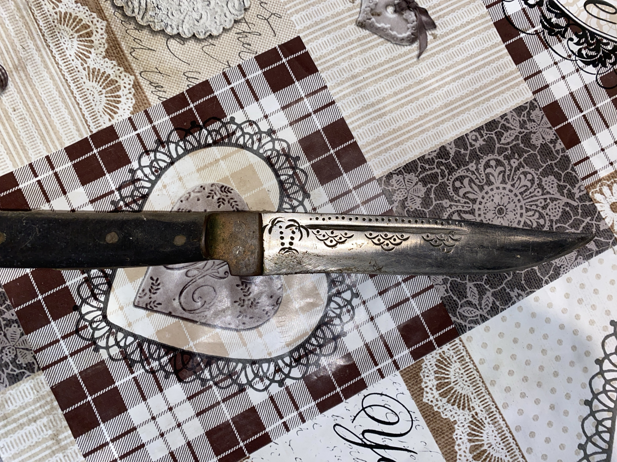 A knife lies horizontally on a patterned surface. The knife has a dark, possibly wooden, handle and a silver-colored blade. The blade is decorated with etched designs. The patterned surface beneath the knife appears to be a collage of different patterns and textures, including a brown and white plaid design, lace, and various floral or decorative motifs. The location and city of Konstantinovo, Bulgaria, are not discernible from the image.