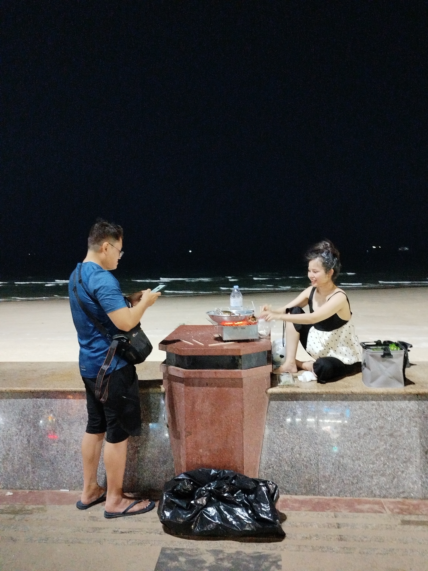 At night in Ho Chi Minh City, Vietnam, a man in a blue shirt and black shorts stands on a low, marble-fronted wall using a cell phone. To his right, a woman in a black and white dress sits on the wall. She is cooking food on a small grill, which is placed on a rectangular brown stone structure. A plastic bottle sits behind the grill. A grey bag is next to the woman. A black plastic bag is on the ground between the man and the brown stone structure. The background shows a dark sky over a beach and ocean. Faint lights are visible on the horizon.