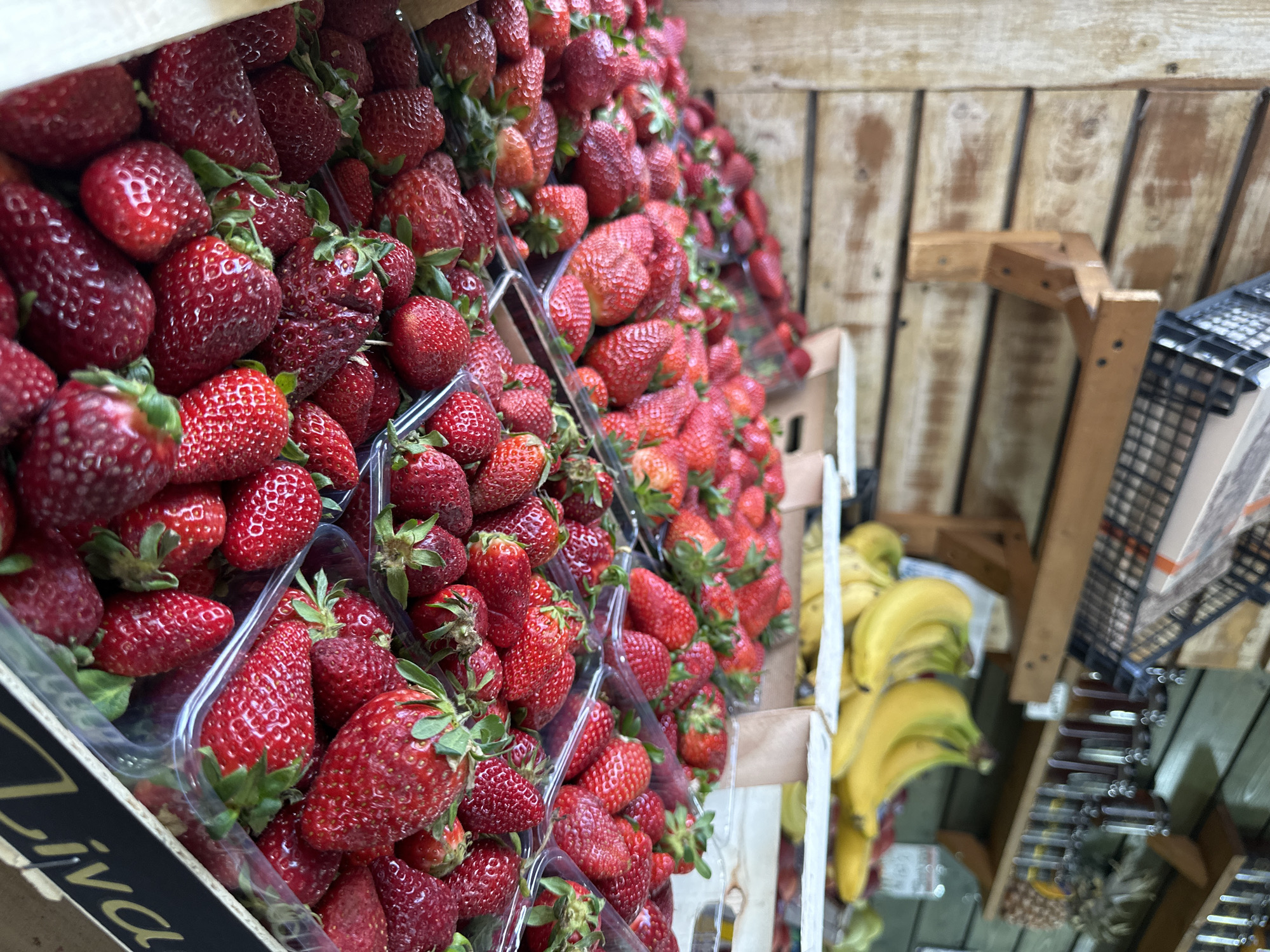 The image shows a display of strawberries for sale, likely in a grocery store or market. The strawberries are arranged in clear plastic containers stacked upon each other. The strawberries are red and ripe. To the right of the strawberries, partially visible, are bananas. The location is likely in Serres, Greece, given the "Liva" text on one of the strawberry boxes. The background appears to be a wooden wall. There are no people visible in the scene.