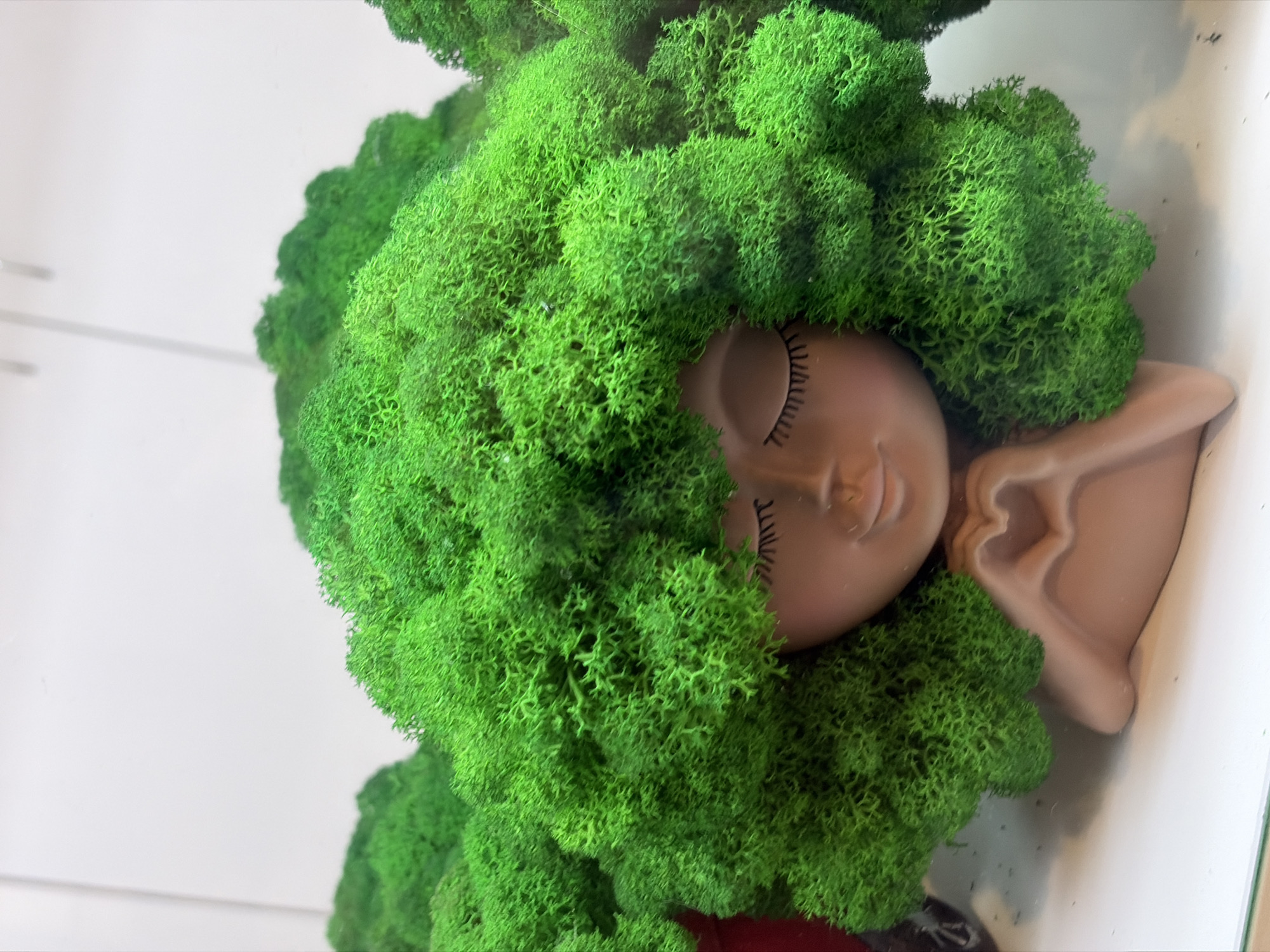 A close-up, eye-level shot focuses on a unique decorative planter. The planter is shaped like the upper torso and head of a person, with a smooth, tan-colored finish. The head is adorned with a voluminous, bright green moss that resembles thick, curly hair. The planter's face is depicted with closed eyes, drawn with thin, black eyelashes, and a gentle, closed-mouth smile. The figure's hands are joined at the chest, forming a heart shape. The planter sits on a white surface, with blurred green mossy shapes in the background, suggesting it's part of a display. The lighting is bright and diffused. The specific location in Saint Petersburg, Russia, is not discernible from the image itself.