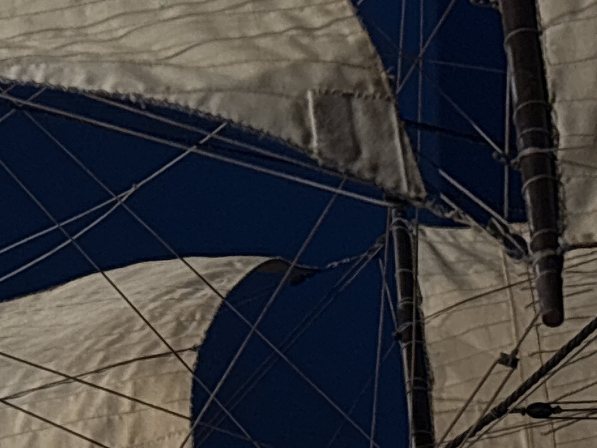 The image shows the sails and rigging of a model ship. The sails are made of white and blue fabric, and the rigging is made of thin strings. The masts are dark brown. The location is not discernible from the image. There is no information available about any people or actions. The environment is unknown.