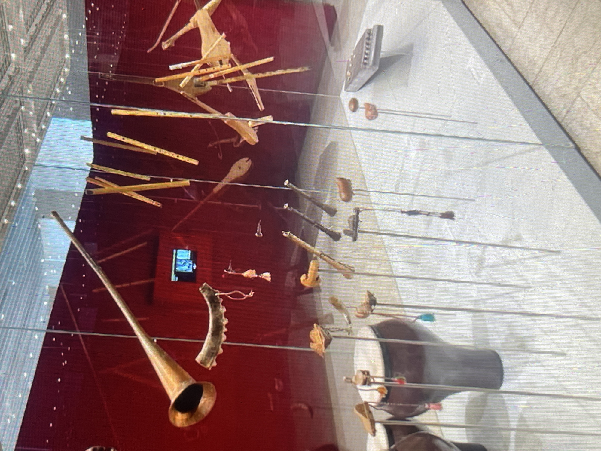 An indoor museum exhibit displays a collection of traditional musical instruments. The instruments are predominantly made of wood and brass, and include various types of flutes, horns, and drums. Some instruments are suspended from thin wires, while others are displayed on stands. The background is a deep red, and the display is encased in glass. The flooring is light-colored and patterned. The exhibit appears to be located in a modern museum. The city of Bishkek, Kyrgyzstan is not mentioned or suggested by any visual cues in the image.