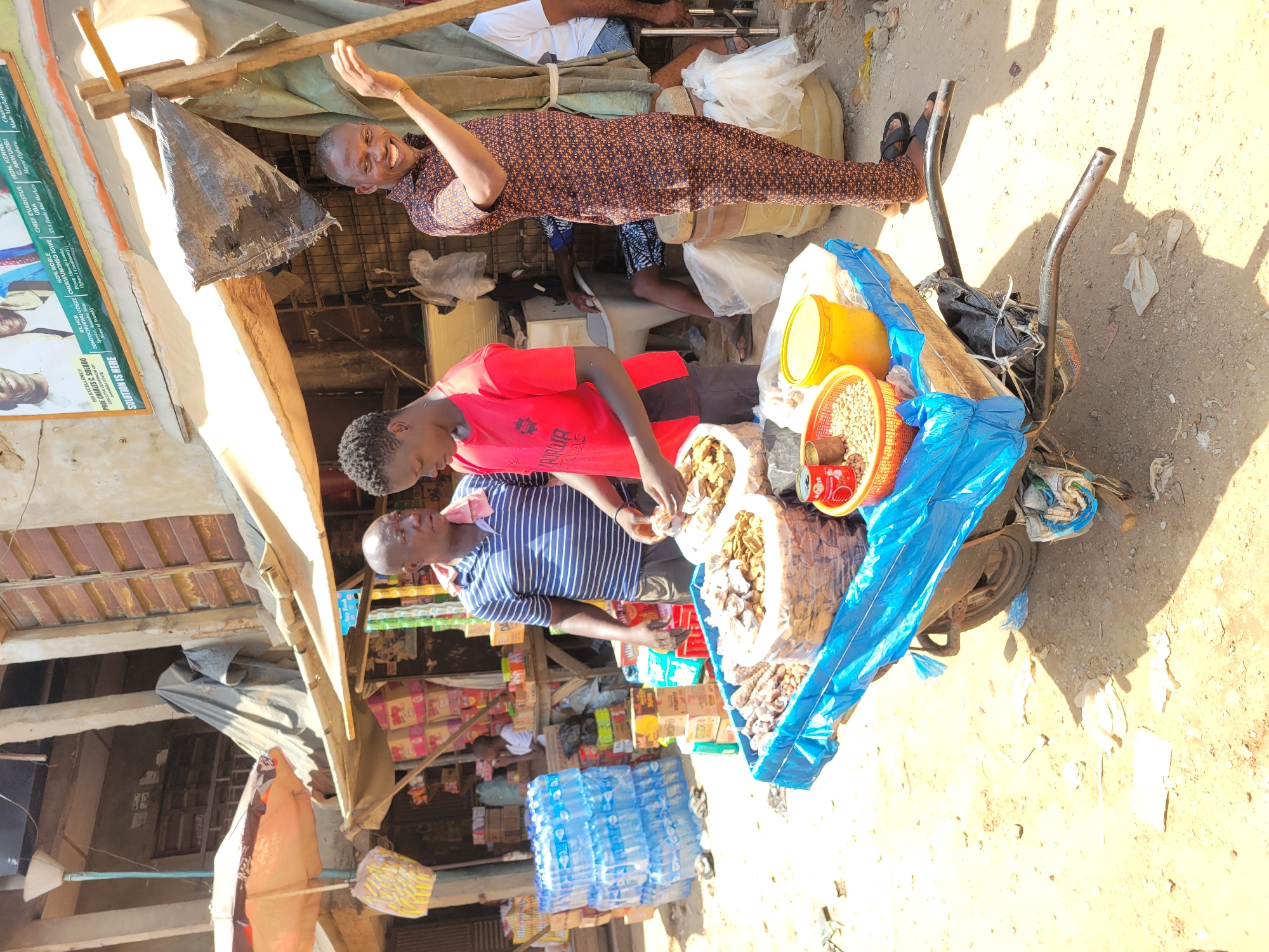 This image depicts a bustling outdoor market scene in Onitsha, Nigeria, captured during daylight hours with bright sun casting strong shadows. The setting features a makeshift stall, characterized by a fabric roof supported by rough wooden beams, providing shade over the goods and vendors. The ground is unpaved, appearing dusty and sandy.
Three main subjects are visible engaging in market activities. A man in a patterned brown and black long-sleeved shirt stands to the right, smiling and raising his right hand, possibly greeting someone or drawing attention. In front of him and slightly to the left, a man wearing a red T-shirt is focused on items displayed on a pushcart or wheelbarrow. Adjacent to him, a man in a blue and white striped shirt also tends to the goods on the cart. Other individuals are partially visible in the background, suggesting a lively environment.
The pushcart holds various merchandise for sale, including several large bags filled with dried goods, potentially nuts, beans, or other staples. A red plastic basket overflowing with what appears to be chickpeas or similar legumes, along with a yellow bucket, are also on the cart, partially covered by blue tarpaulin. In the background, numerous crates of bottled water are stacked, indicating a broader range of products available.
A notable detail is a large political poster affixed to the left side of the stall. Prominent text on the poster reads "SOLUTION IS HERE" and features names and titles such as "HIS EXCELLENCY PROF CHARLES C. SOLUDO EXECUTIVE GOVERNOR OF ANAMBRA STATE," "RT. HON SOMTOCHUKWU UDEZE Speaker member State House of Assembly," "CHIEF EVARISTUS UBA SSA Trade And Markets Major Ogbaru," "HON. KENI C. AKWUOBI Hon. Noble (Ogbara Constituency)," and "HON. NOBLE CHUKWUNONSO IGWE Deputy Minority Leader Ogbara Constituency." This text provides specific location cues within Anambra State, Nigeria. The overall atmosphere conveys the vibrant, everyday commerce of a local market.