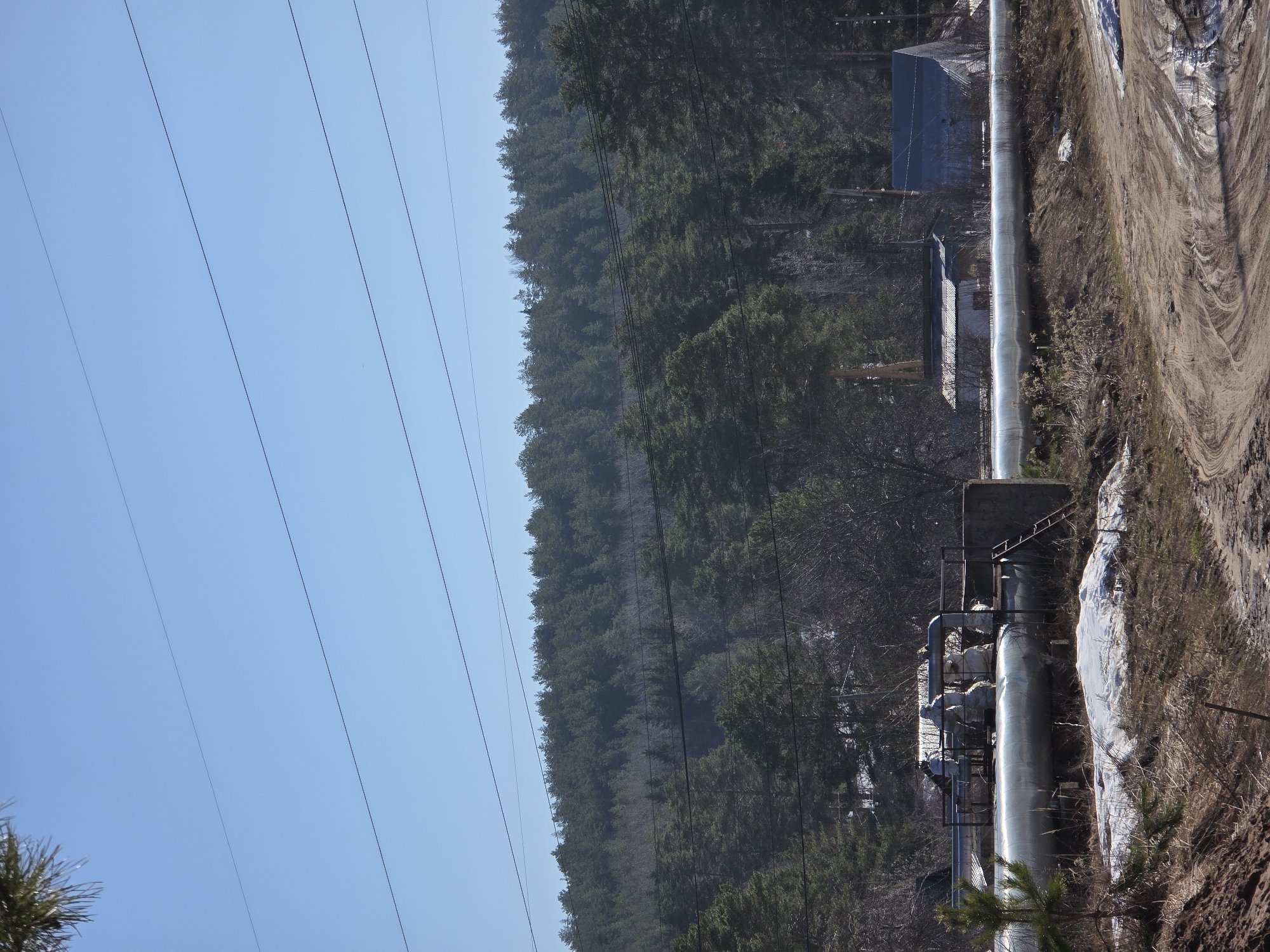 The image depicts an industrial scene in Chaikovsky, Russia. A large, silver-colored pipeline runs through a wooded area, parallel to a dirt road. The pipeline is elevated on concrete supports and has attached machinery. Power lines are visible against a clear blue sky on the left side of the image, near a forest on the ridge. Two small buildings are near the pipeline. The ground is covered with dirt, vegetation, and patches of snow.