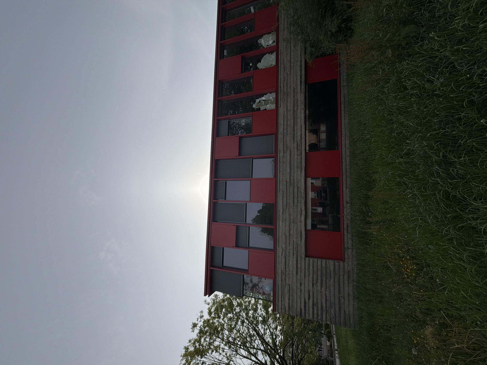The image depicts a tall, multi-story building with a red and grey facade. The building features a grid-like pattern of windows and solid panels. A section of the building's facade is textured with vertical lines, possibly concrete or wood. Vegetation, including grass and bushes, grows along the base of the building. A tree is visible in the lower left corner. The sky is overcast, with the sun appearing as a bright, diffuse light source. The photograph is taken from a low angle, looking upwards at the building. The location appears to be Esch-sur-Alzette, Luxembourg, based on the architectural style and vegetation.