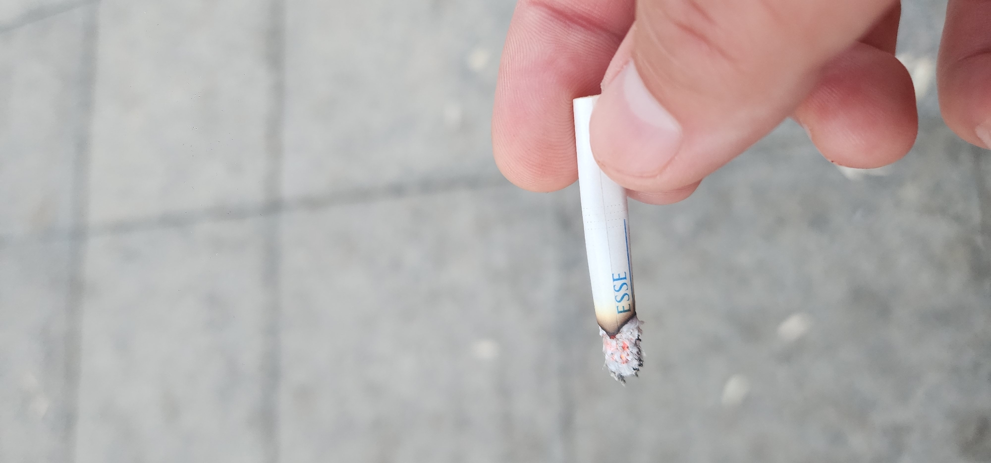 In Türkmenbaşy, Turkmenistan, a person's fingers hold a lit cigarette with the brand name "ESSE" visible on its white filter. The cigarette is smoldering at the tip, with ash and glowing embers present. The background is a blurred, gray surface that appears to be pavement or a similar textured outdoor ground. The lighting suggests it is daytime.
