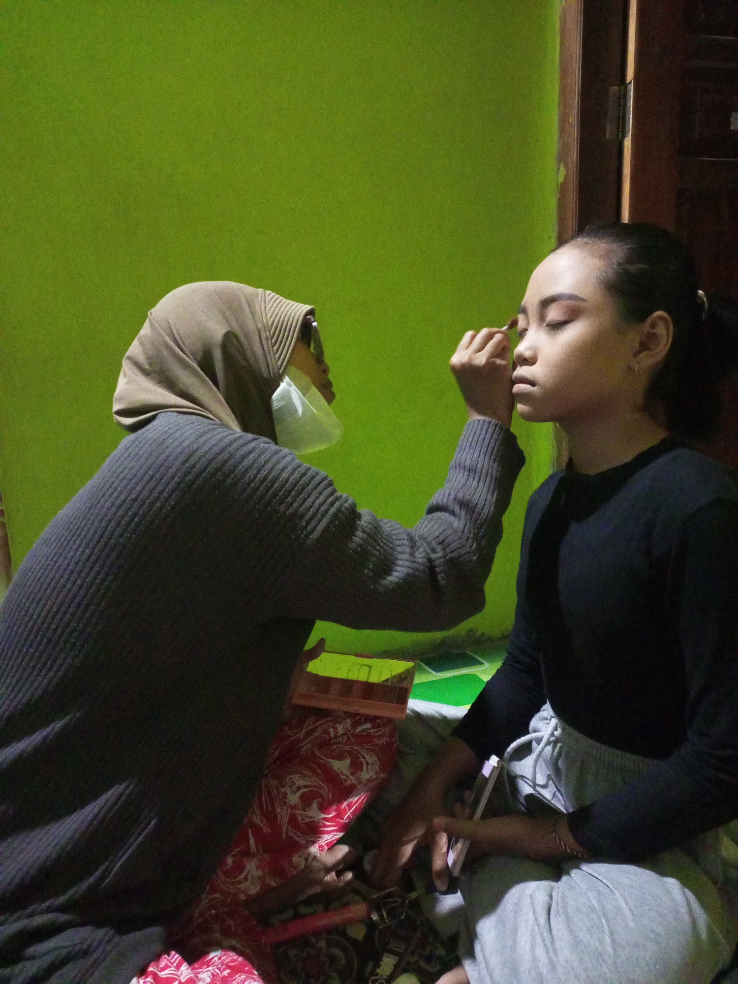 In Nanggulan, Indonesia, an individual wearing a brown hijab and a face mask is applying makeup to another individual. The person receiving makeup is seated and facing towards the right of the frame, with their eyes closed. The makeup artist is using a brush on the recipient's eyelid. The background features a bright green wall and a wooden doorframe. The floor is covered with patterned fabric and a small wooden box. Both individuals are dressed casually, with the makeup artist in a dark ribbed sweater and the recipient in a black long-sleeved shirt and grey sweatpants.