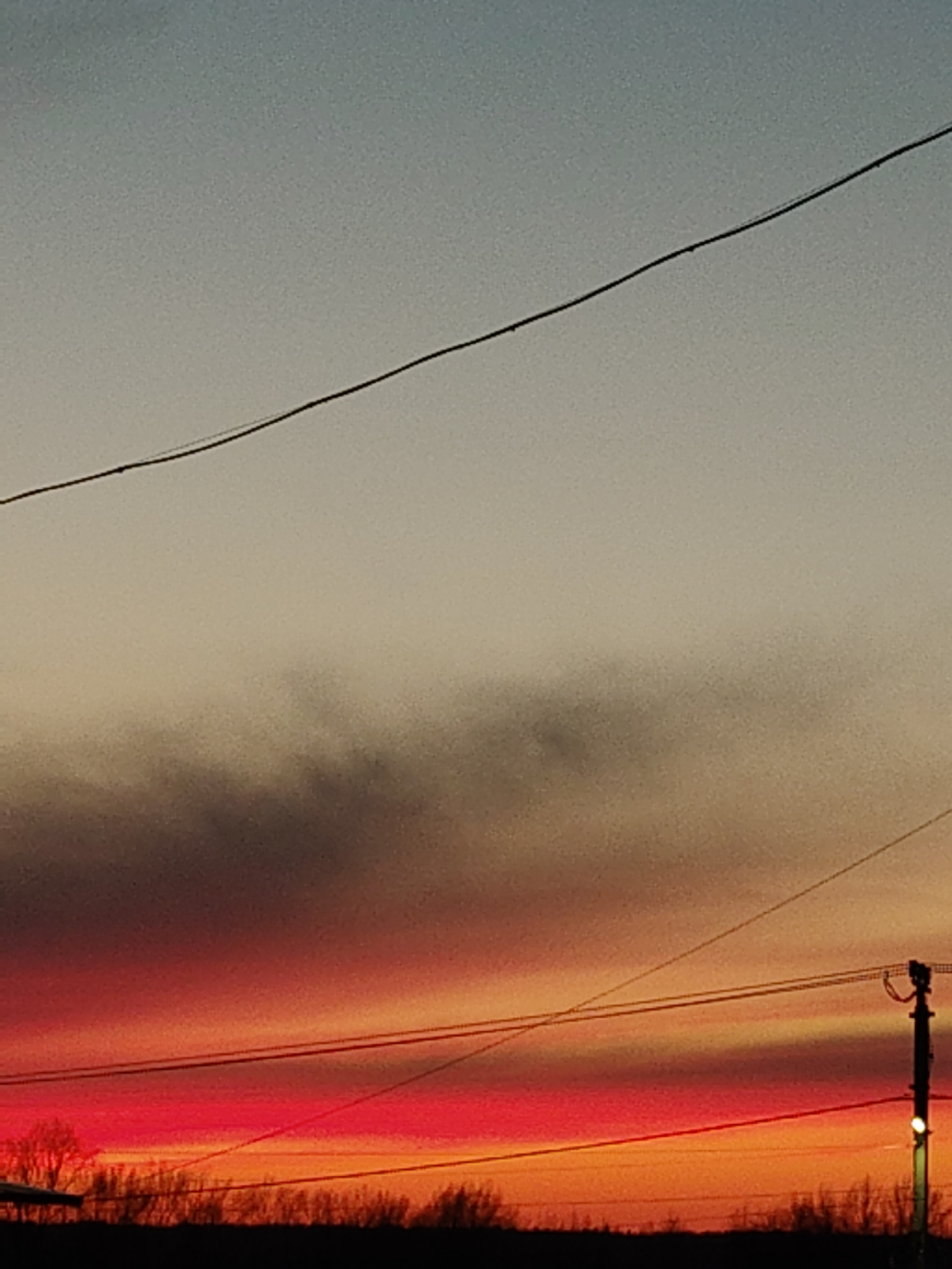 The image depicts a sky at sunset with a gradient of colors ranging from red and orange near the horizon to grey and pale blue higher up. Silhouetted trees and vegetation are visible along the bottom edge of the frame, forming a dark, irregular line. Several power lines are strung across the sky, and an electrical pole is visible on the right side, with a light glowing. The location is unknown. There are no people visible in the image.