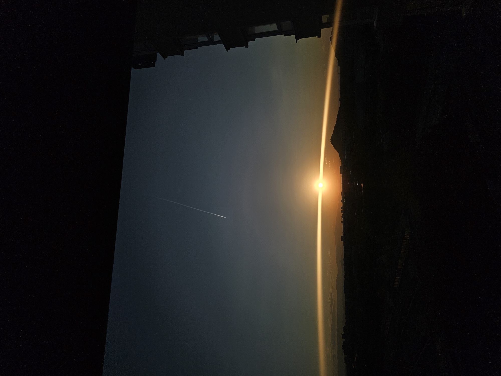 The image depicts a cityscape with a bright sun emitting a vertical ray of light. A contrail cuts across the upper left portion of the frame. The view is likely from an elevated position, possibly a building in Plovdiv, Bulgaria, as suggested by the cityscape. Buildings are silhouetted against the light on the right, with a darker block framing the image on the left and across the top. The image appears to be taken during sunrise or sunset.