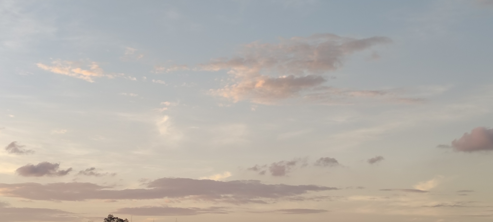 The image shows a sky with various cloud formations. The color palette is pastel, with hues of light blue, pale pink, and soft orange, suggesting either dawn or dusk. The clouds are scattered and varied in shape, some appearing thin and wispy while others are more dense and defined. The scene is devoid of any visible human presence or man-made structures, implying a wide-angle view focusing on the atmospheric conditions. Given the specification that the image is located in Lhokseumawe, Indonesia, the atmospheric conditions suggest a tropical or subtropical environment.