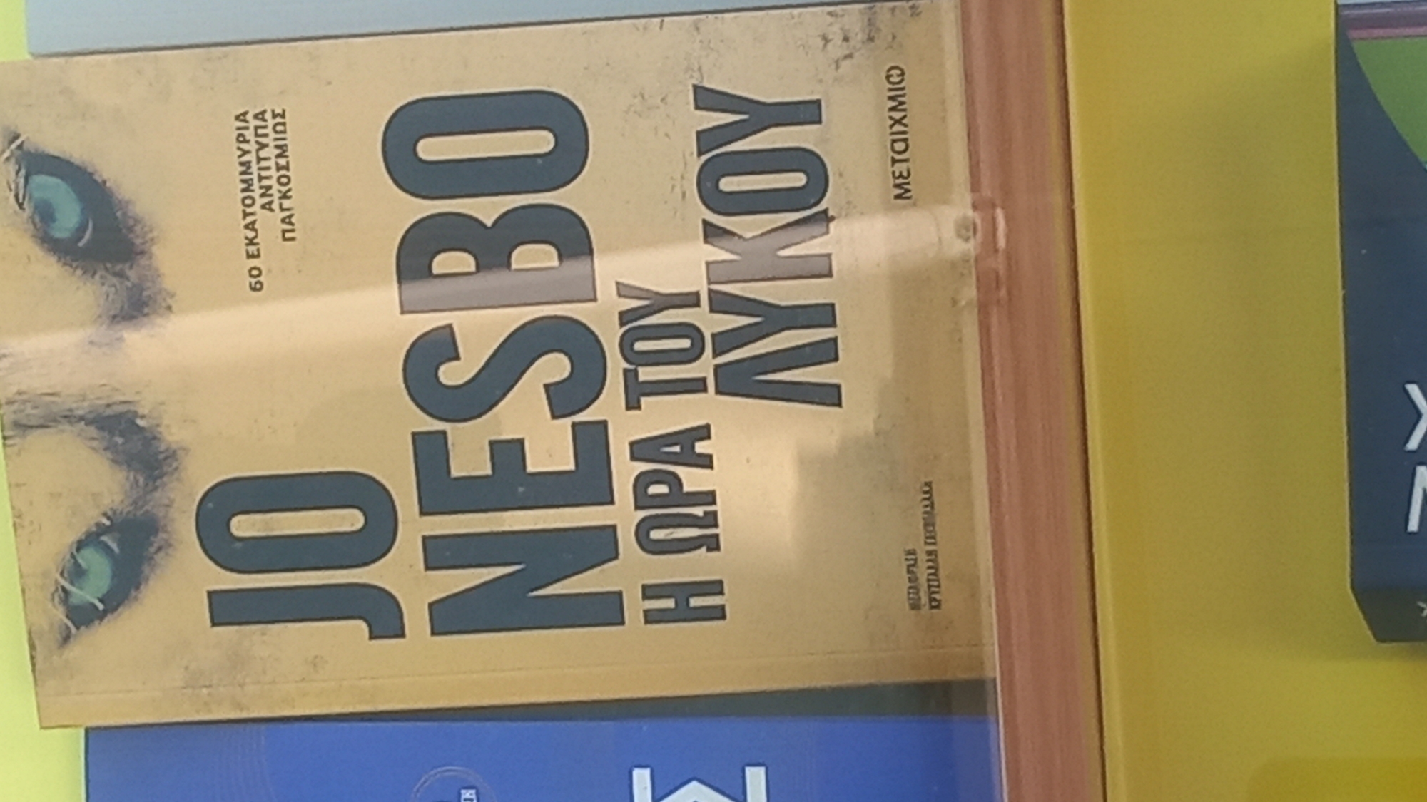 The image shows a book cover with the title "JO NESBO H ORA TOY LYKOU" in large, bold, dark blue letters against a yellow background. The book cover also features a close-up image of a wolf's eyes. In smaller text on the right side of the cover, it reads "60 EKATOMMYRIA ANTITYPA PANKOSMIOΣ". At the bottom left, there is smaller text that reads "METAFIBH KPYSTALLAH EKEYALLACH". On the bottom right, also in smaller text, it says "METAIXMIO". The book is placed on a shelf in a store. The surrounding environment is out of focus but appears to be a retail setting.