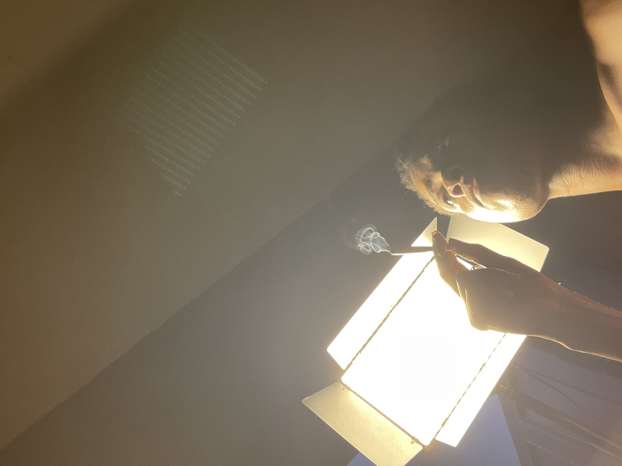 An upward-angled, low-light photograph shows a partially visible person holding a lit cigarette. Smoke emanates from the tip of the cigarette. The person's face, neck, and arm are partially illuminated by a bright light panel positioned in the lower center of the frame. The background is a dark, slightly textured wall. There are no clear visual markers suggesting the location of Onitsha, Nigeria.