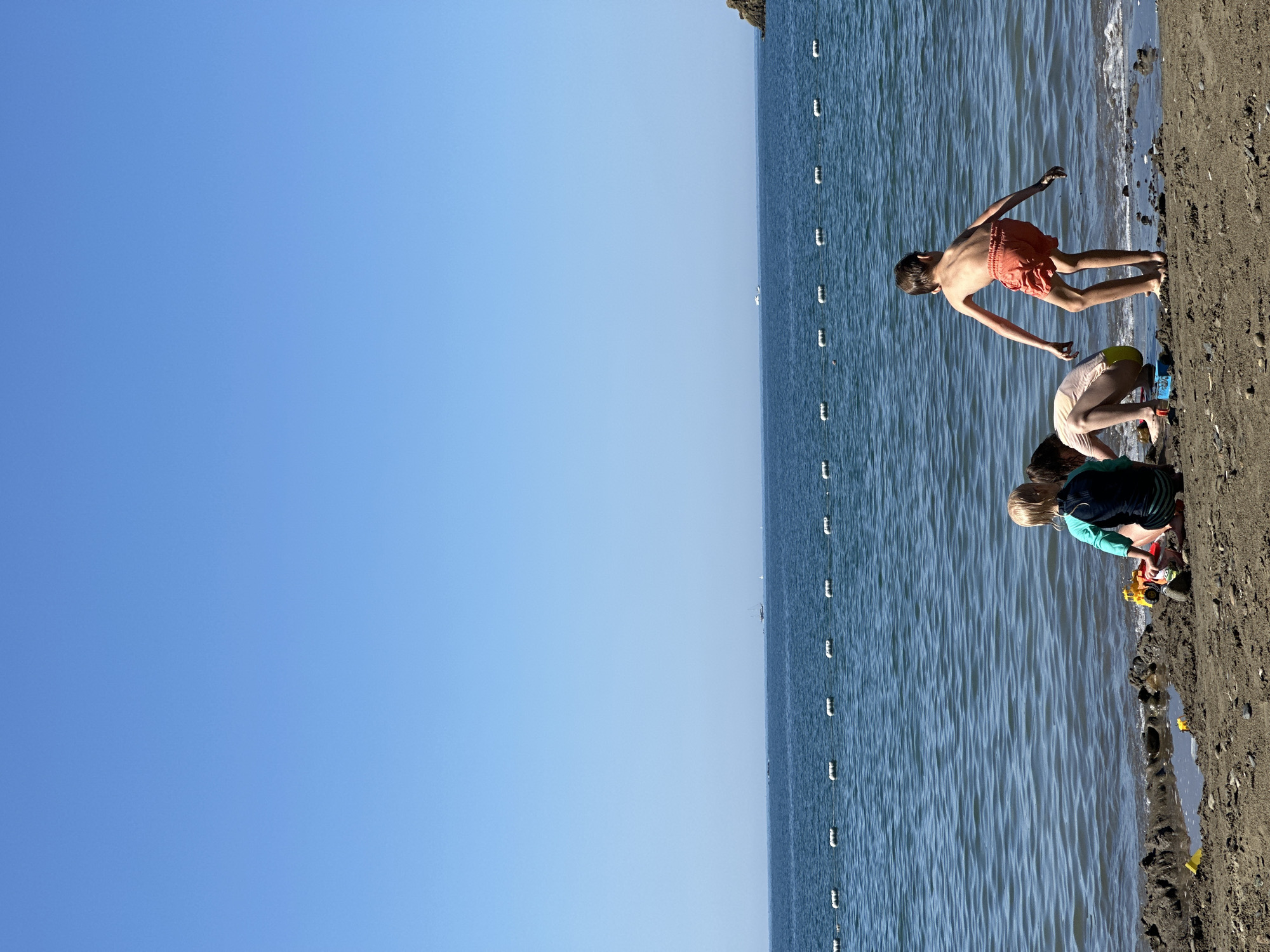 The image shows a beach scene in Fethiye, Turkey, with children playing at the water's edge. The sky is clear blue, and the sea is a deep blue color with a line of white buoys. Three children are on the beach, one is shirtless and wearing orange shorts, bending over with arms extended; another is wearing light-colored shorts, and a third is wearing a blue and green top. They appear to be playing in the sand near the water. The beach sand is dark. The perspective of the image is rotated 90 degrees clockwise.