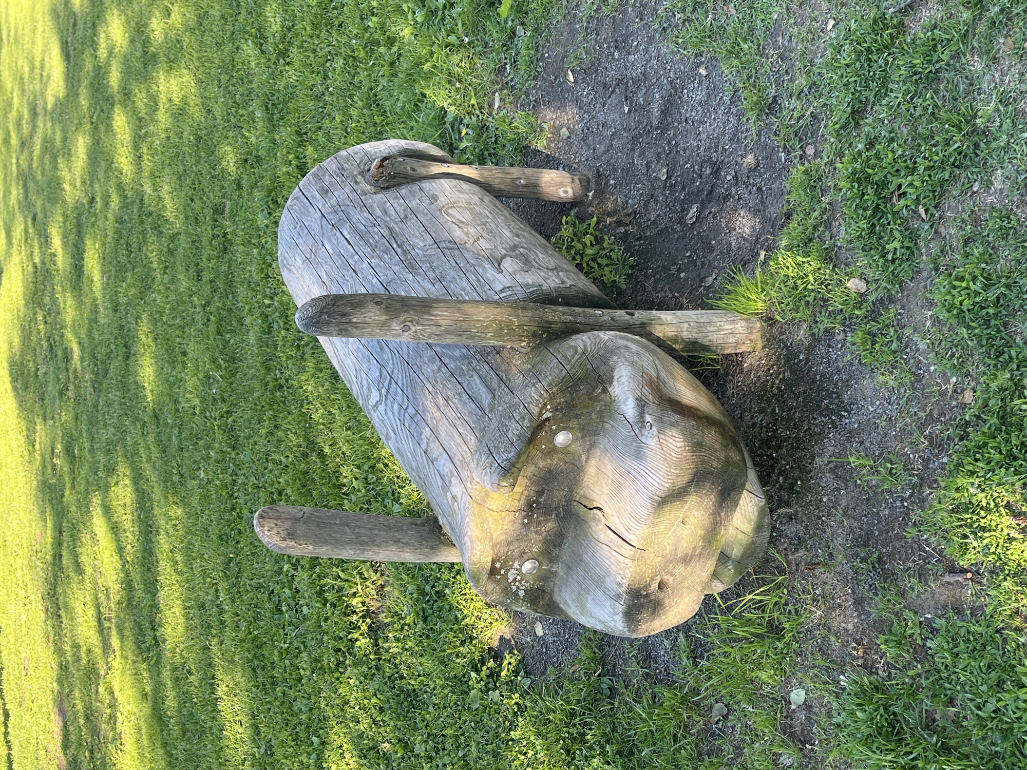 The image shows a wooden sculpture of a pig in a park. The sculpture is made from a log and has two wooden posts for legs at the front and two at the back. It is positioned on a grassy area with some gravelly patches. The lighting suggests it is daytime. This sculpture is located at Tuiranpuisto park in Oulu, Finland.