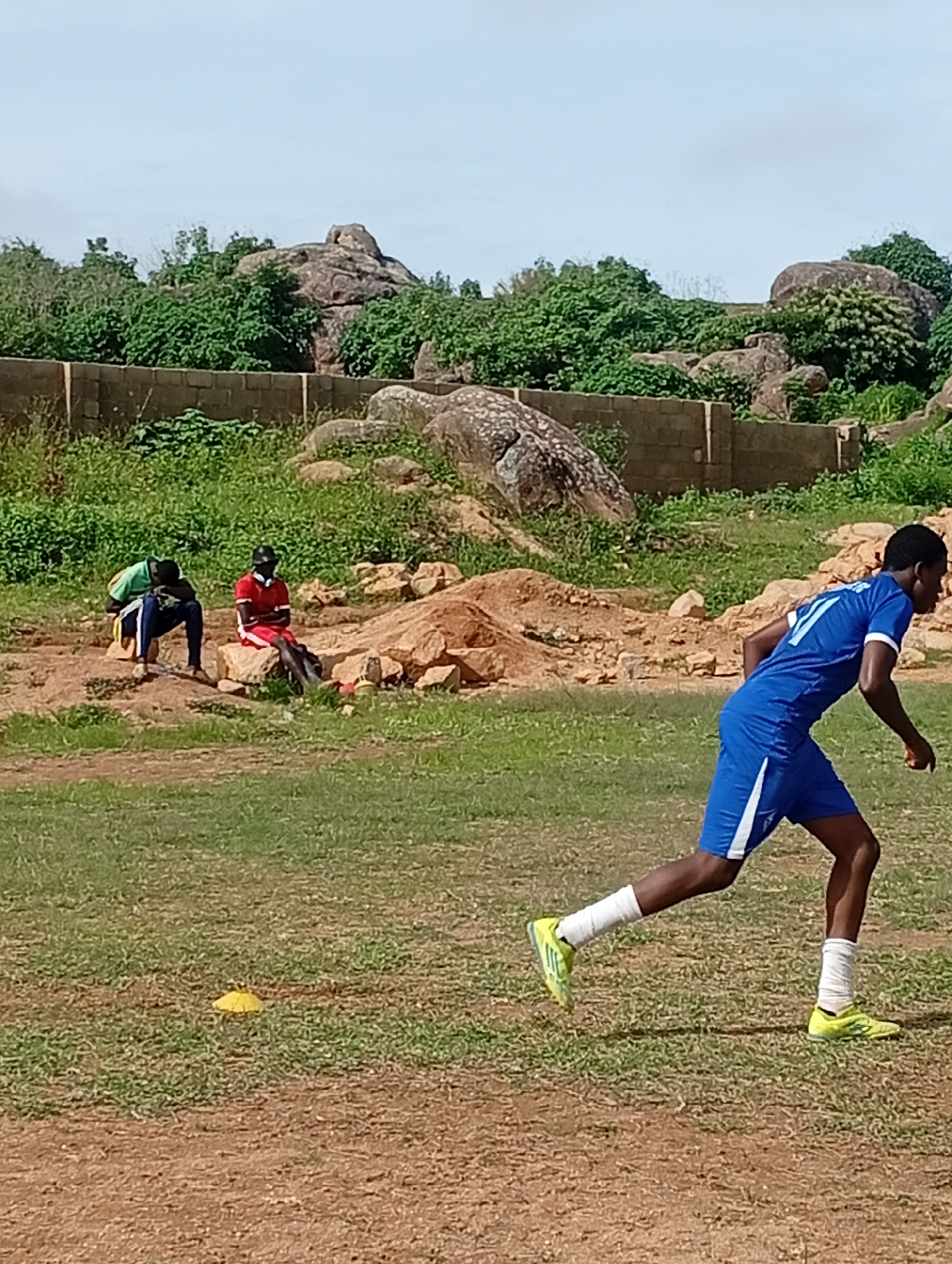 A young male soccer player, wearing a blue jersey with white trim and the number 11, blue shorts with white trim, white socks, and yellow cleats, runs across a grassy field. In the background, two other young males sit on rocks. One, in a red jersey and shorts, sits with his arms crossed. The other, in a green shirt and blue pants, is bent over with his head in his hands. The field is dusty with patches of dry grass. In the distance, a stone wall encloses a rocky, vegetated area with large boulders. This scene is consistent with an outdoor sports training ground in the city of Jos, Nigeria, which is known for its rocky terrain and numerous boulders.