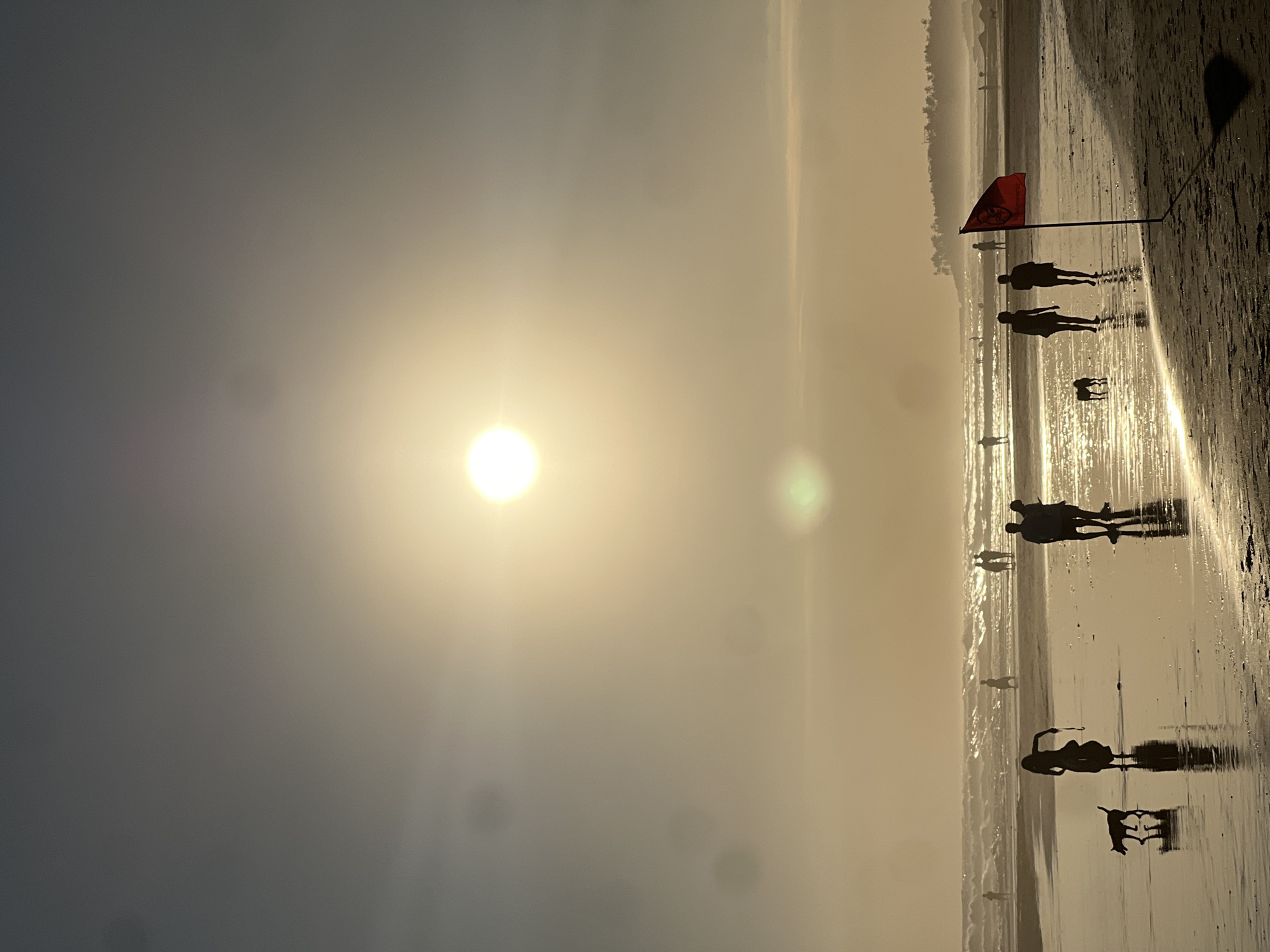 A vertical, silhouette photograph captures a beach scene at sunset in North Kuta, Indonesia. The sun, a bright, hazy orb, dominates the upper half of the frame, casting a warm, golden light across the scene. The sky transitions from a pale grey at the top to a soft, hazy yellow near the horizon. In the middle ground, gentle waves roll onto the shore, their crests catching the light. Several figures are visible on the wet sand, appearing as dark silhouettes against the bright water and sky. On the left, a person walks with a dog. Further back, a group of people stand together, and another individual walks near the water's edge. On the right side of the frame, two figures walk away from the camera, and in front of them, a red flag with a dark symbol is planted in the sand, casting a long shadow. The sand itself is wet and reflective, mirroring the figures and the sky. In the far background, a line of trees can be seen along the distant coastline. The overall atmosphere is serene and tranquil, evoking the end of a day at the beach.