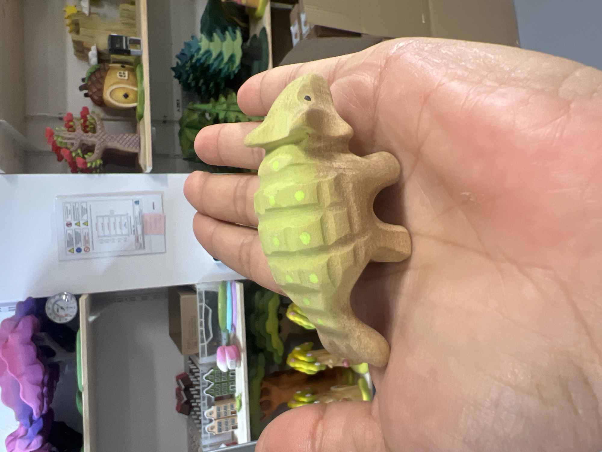 A person's hand is holding a small, light green, wooden toy dinosaur with yellow dots and a carved texture. The dinosaur is positioned on the palm, facing to the right. The background shows shelves filled with miniature toys and decorations, including houses, trees, and a clock. The environment appears to be a room with shelving units.